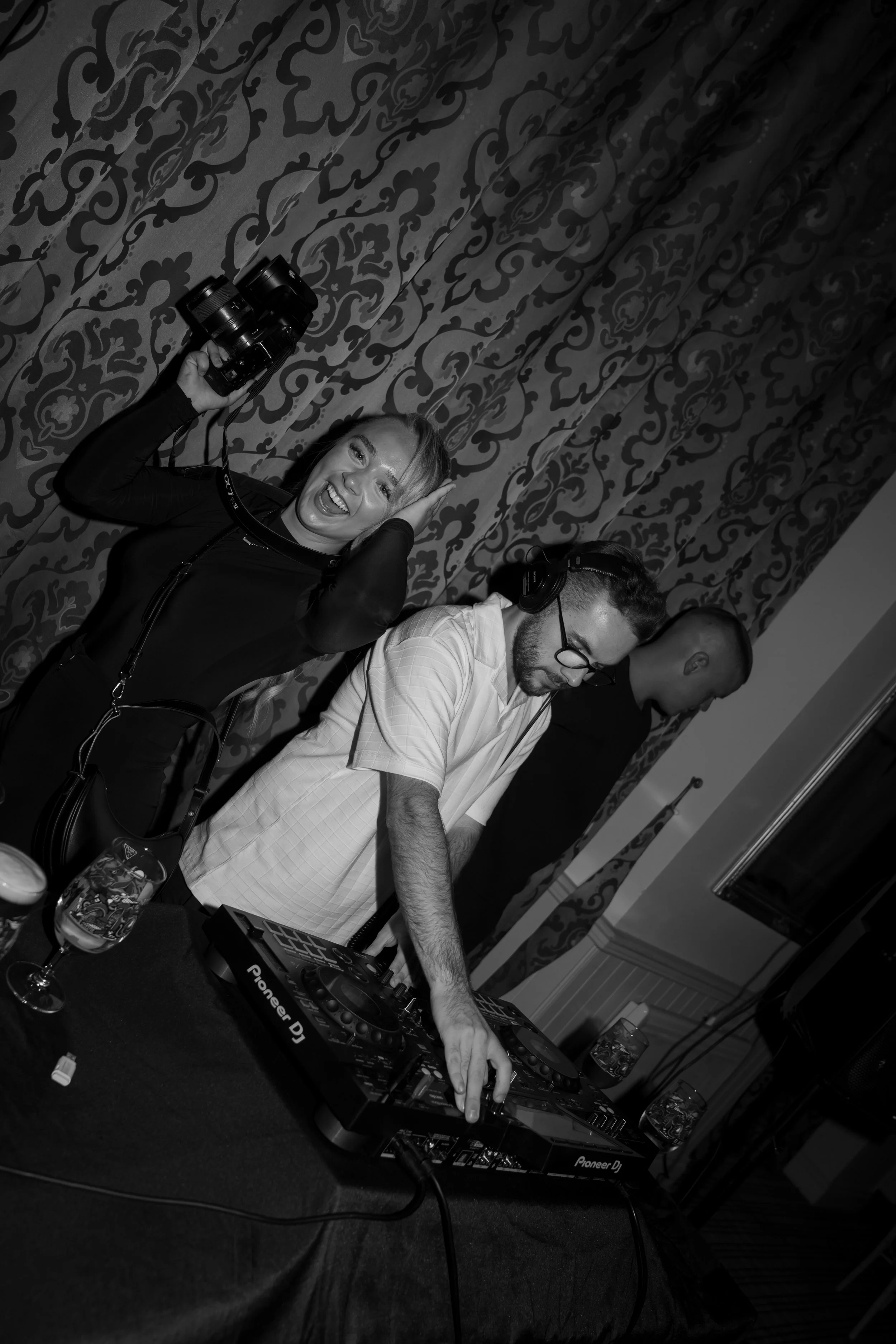 A woman holding a camera smiling excitedly, a DJ with glasses mixing music at a DJ console, and a man in the background, all in a room with ornate patterned curtains.