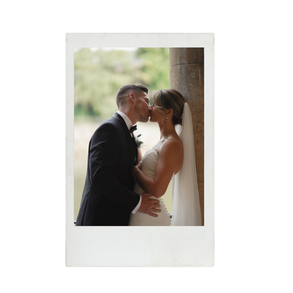 A newlywed couple, dressed in wedding attire, sharing a kiss indoors near a column with nature visible in the background.