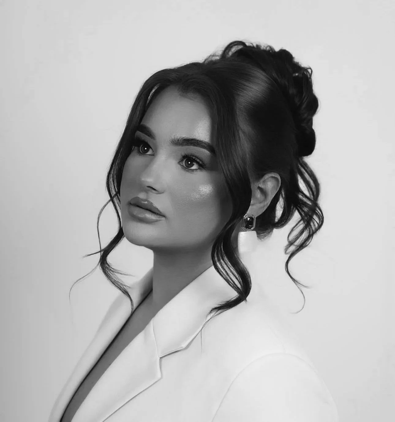 Black and white portrait of a woman with styled hair, makeup, earrings, and a white blazer, against a plain background.