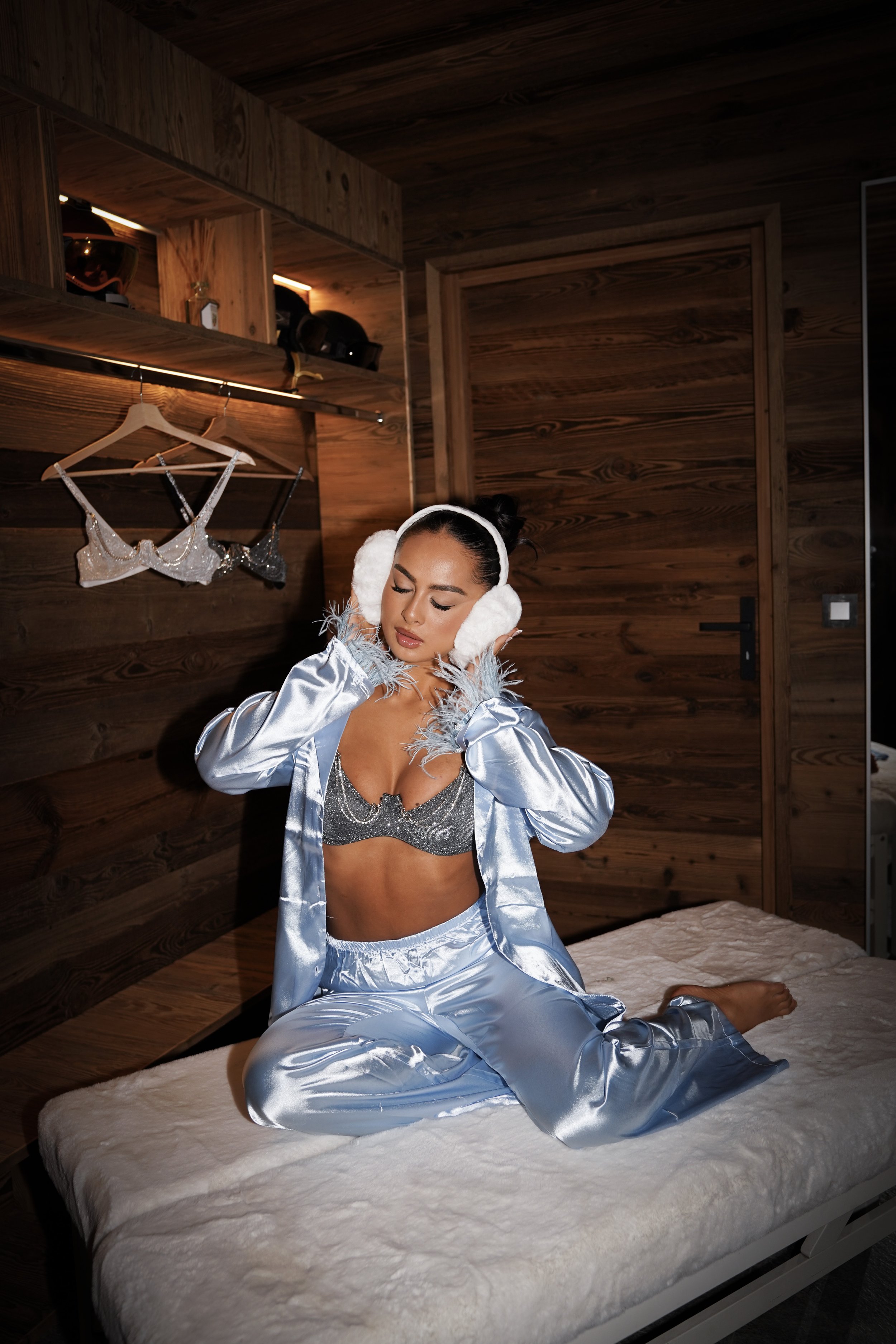 Woman wearing silver satin pajamas and earmuffs, sitting on a bed with a wooden closet in the background, in a cozy room.