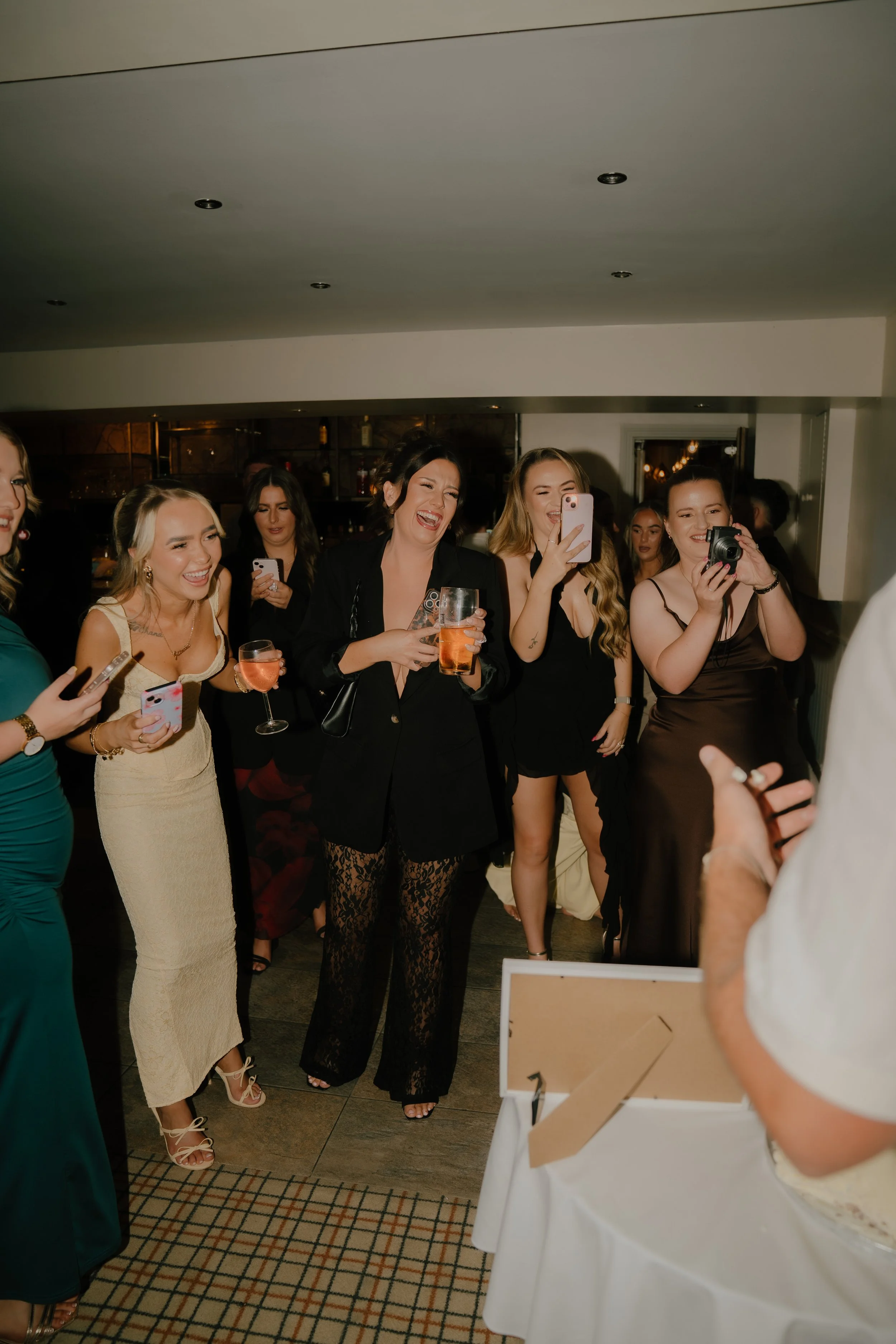 A group of women are smiling, laughing, and taking photos at a celebration, with some holding drinks, in a warmly lit indoor setting.