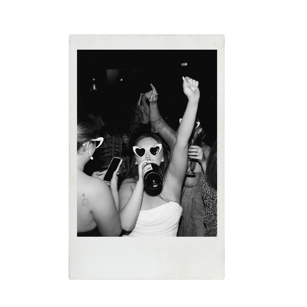 People partying and dancing at a social event, wearing sunglasses and holding drinks, with a woman drinking from a bottle and wearing heart-shaped sunglasses, captured in a black and white Polaroid-style photo.