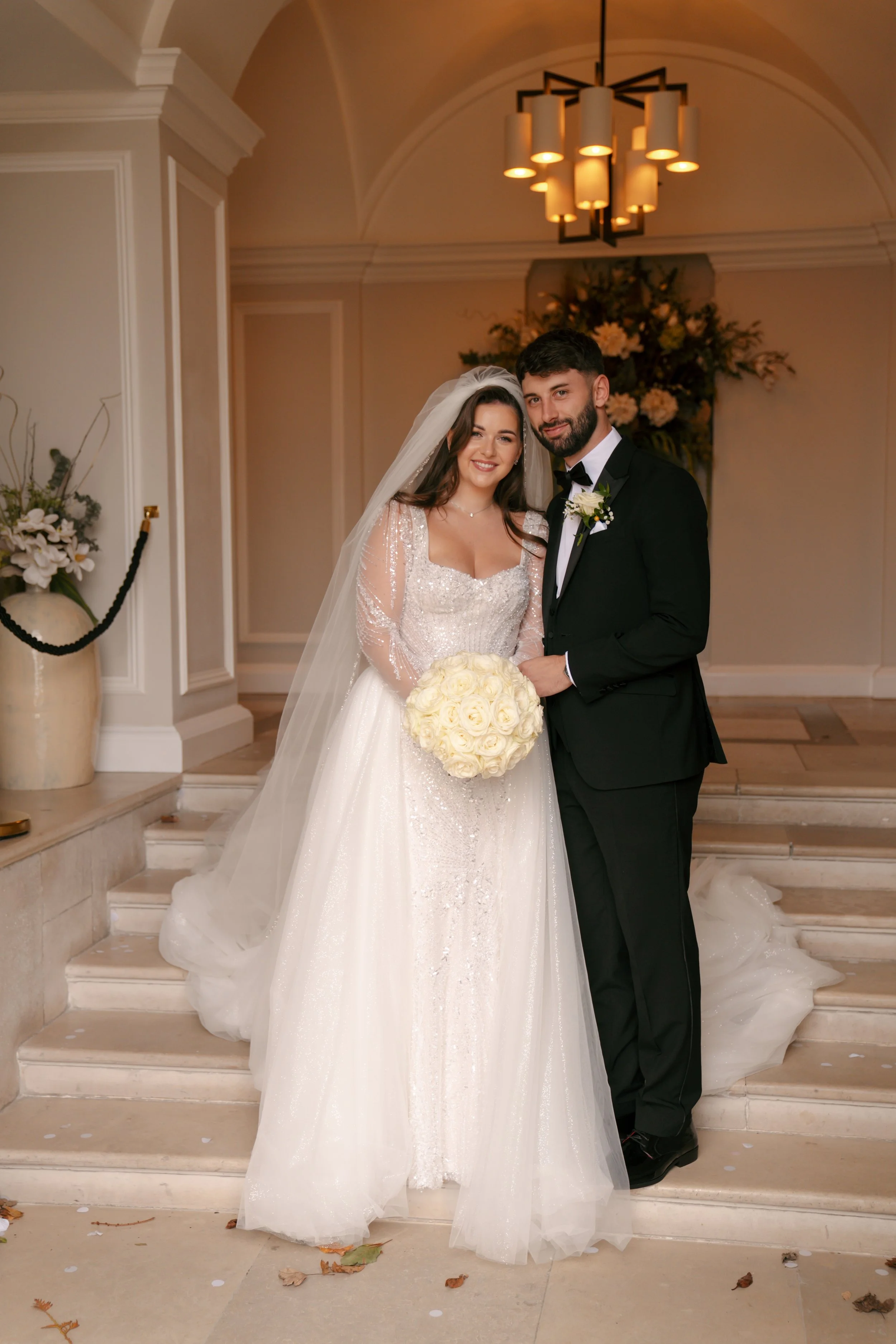 A bride and groom standing together on the steps of a wedding venue, smiling at the camera. The bride is in a white wedding dress with long sleeves and holding a bouquet of white roses, while the groom is in a black tuxedo with a bow tie and boutonnière. The background features a floral arrangement and a modern chandelier.