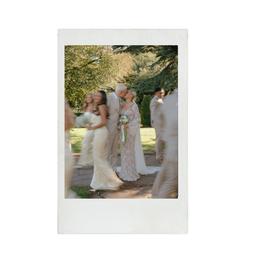 A bride and groom kiss outdoors surrounded by wedding guests, with trees and greenery in the background.