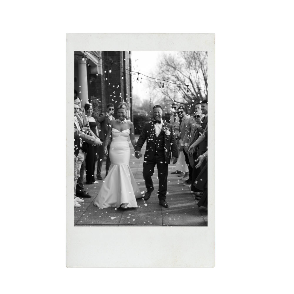 Black and white photo of a newlywed couple walking outdoors with friends and family throwing confetti.