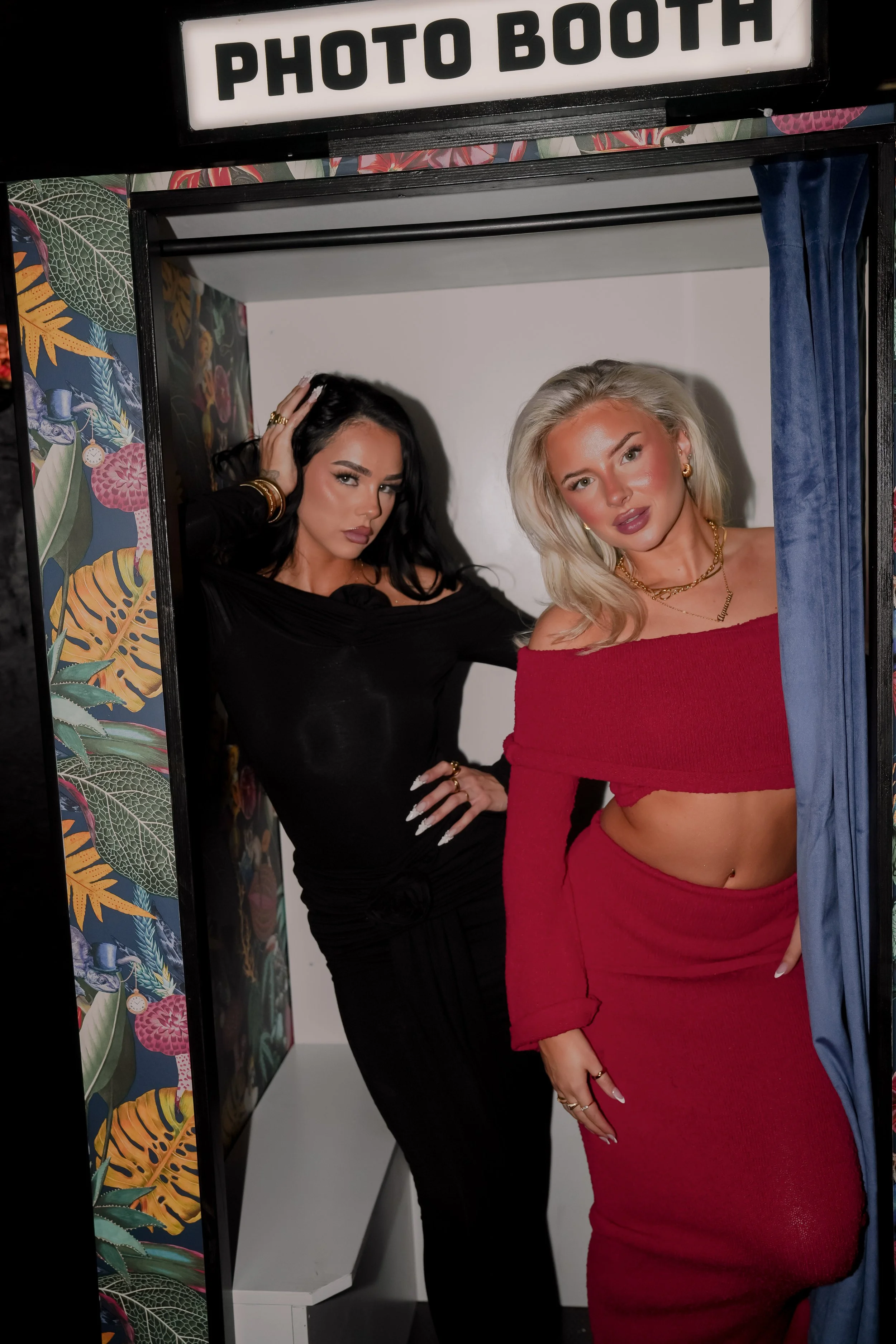 Two women posing in a photo booth, one in a black dress and the other in a red off-the-shoulder outfit with a cutout, inside a decorated booth with a sign that says 'Photo Booth' at the top.