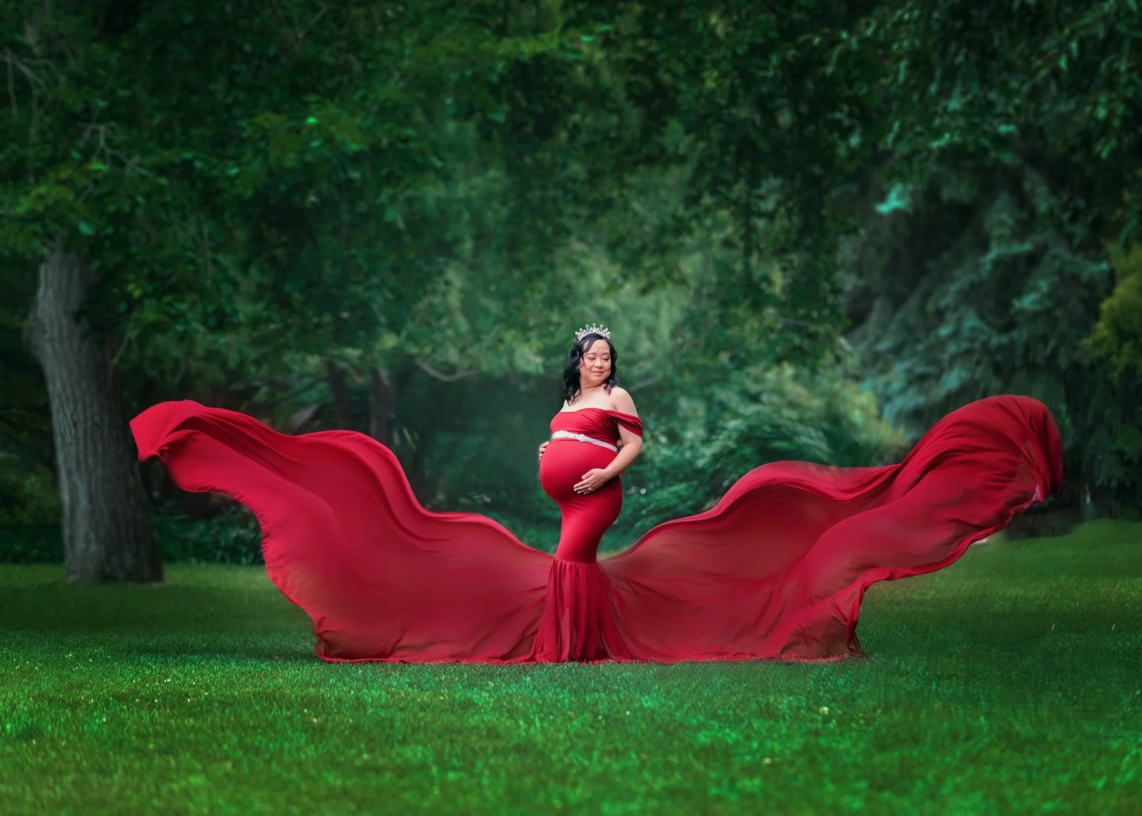 A pregnant woman in a red gown with flowing fabric standing on a lush green lawn in a forested area, wearing a tiara and holding her belly.
