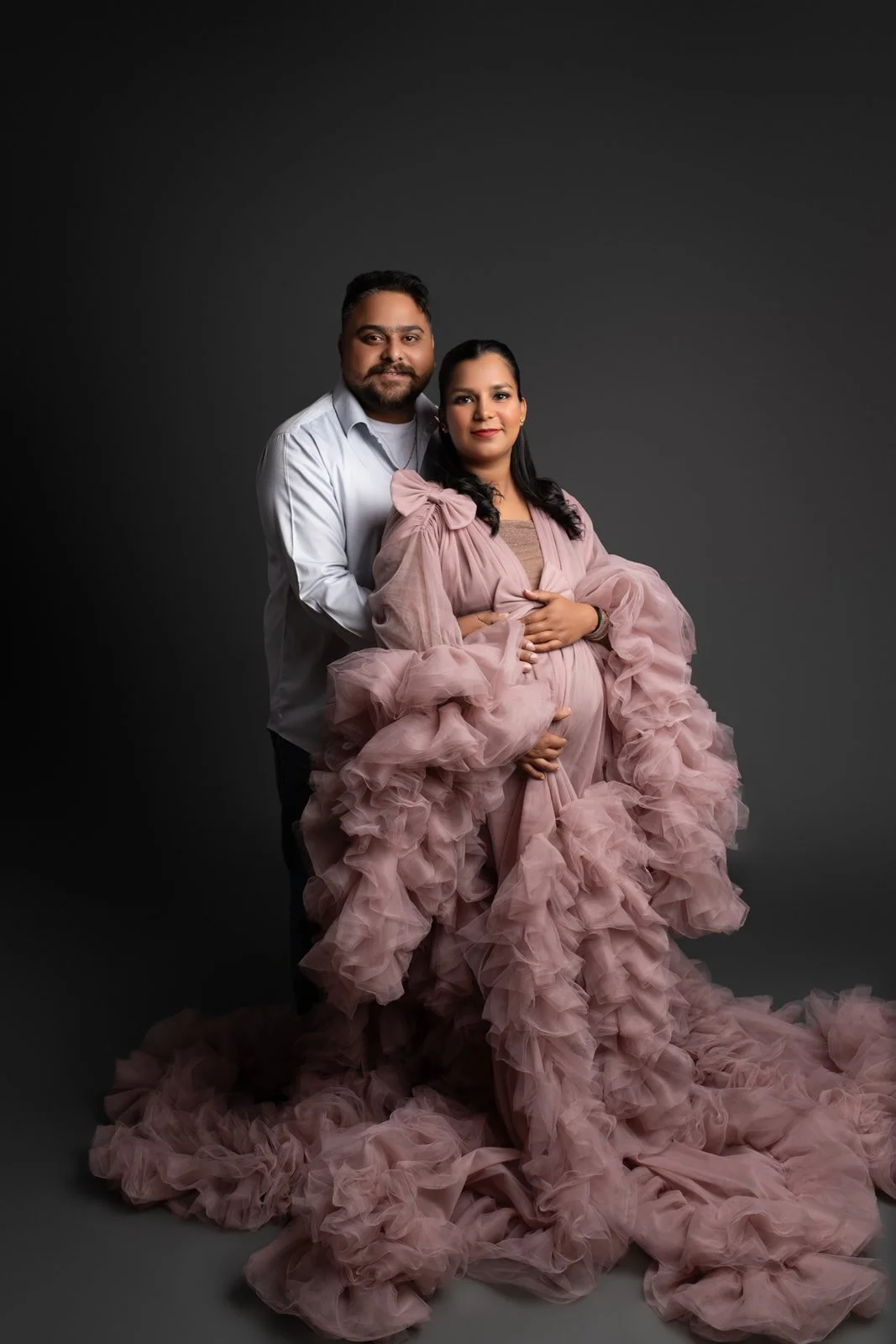 A couple, the woman in a flowing pink ruffled gown and the man in a white shirt, poses together against a dark gray background.