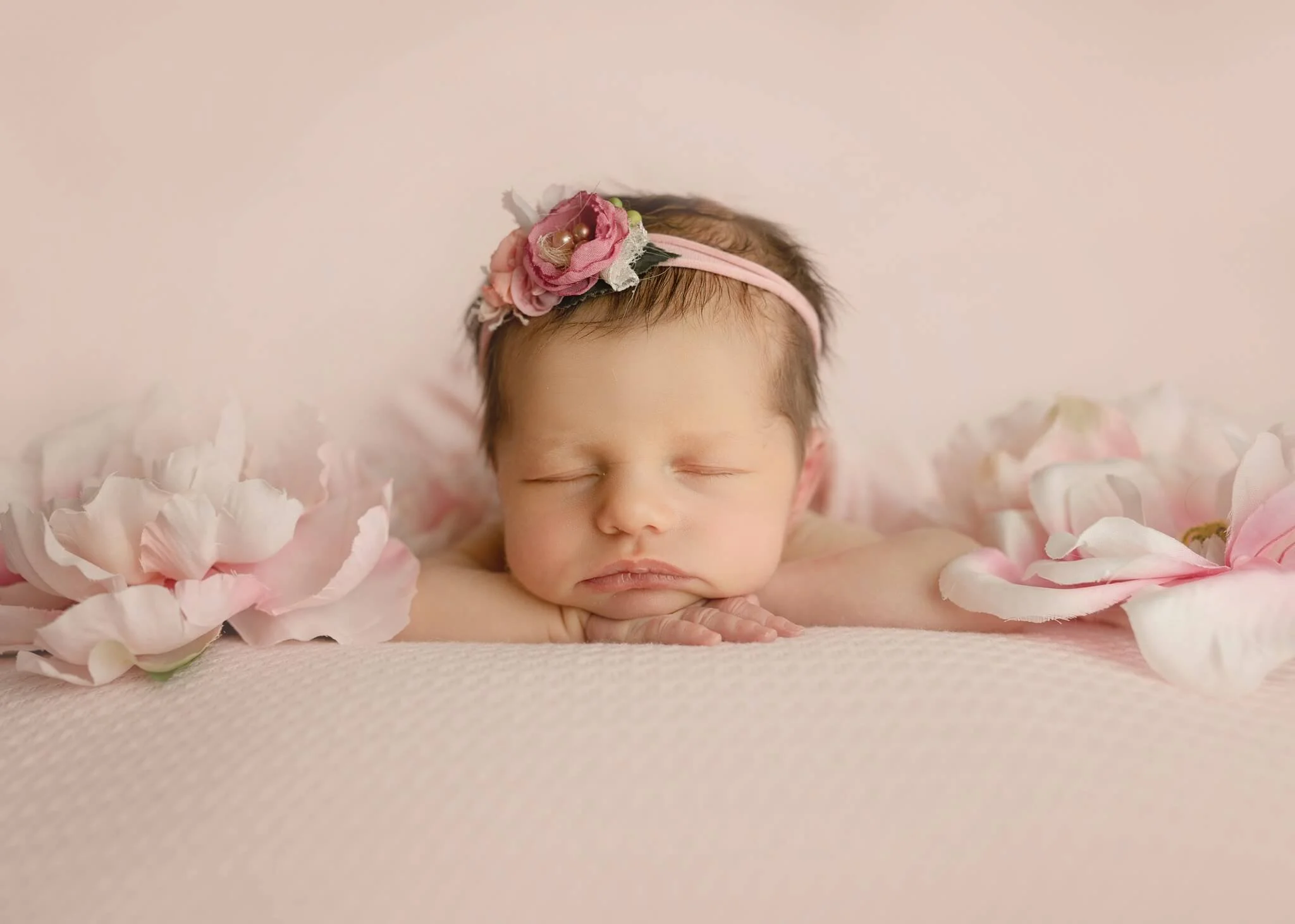 Sleeping newborn baby with a pink floral headband, surrounded by pink and white flowers, lying on a soft pink surface.