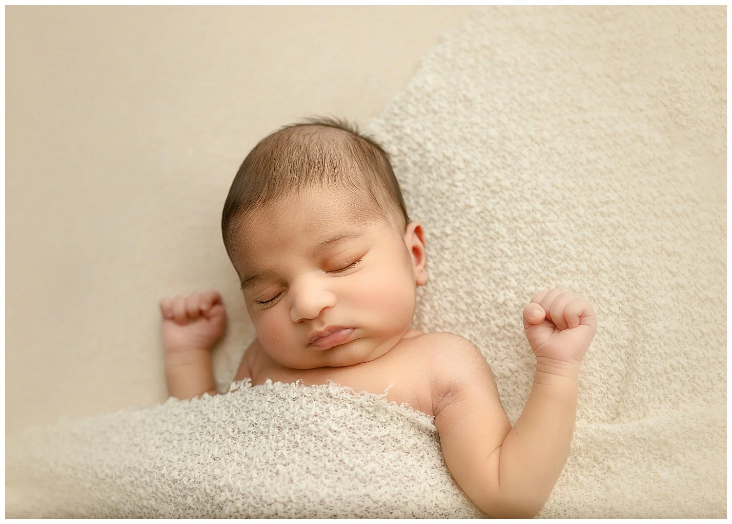 Close-up of a sleeping baby with light skin, dark hair, lying on a soft textured blanket, with fists clenched