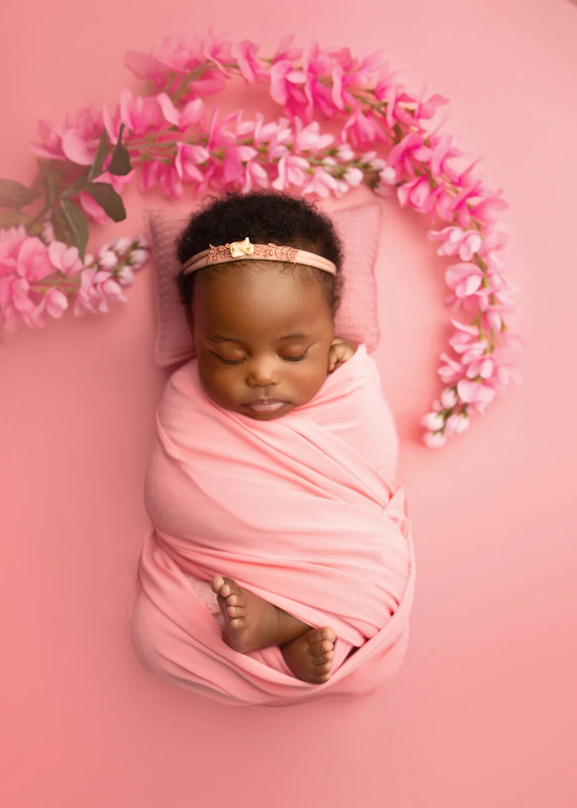 A sleeping baby girl wrapped in a pink blanket, lying on a pink surface with pink peonies arranged in a circle around her head.