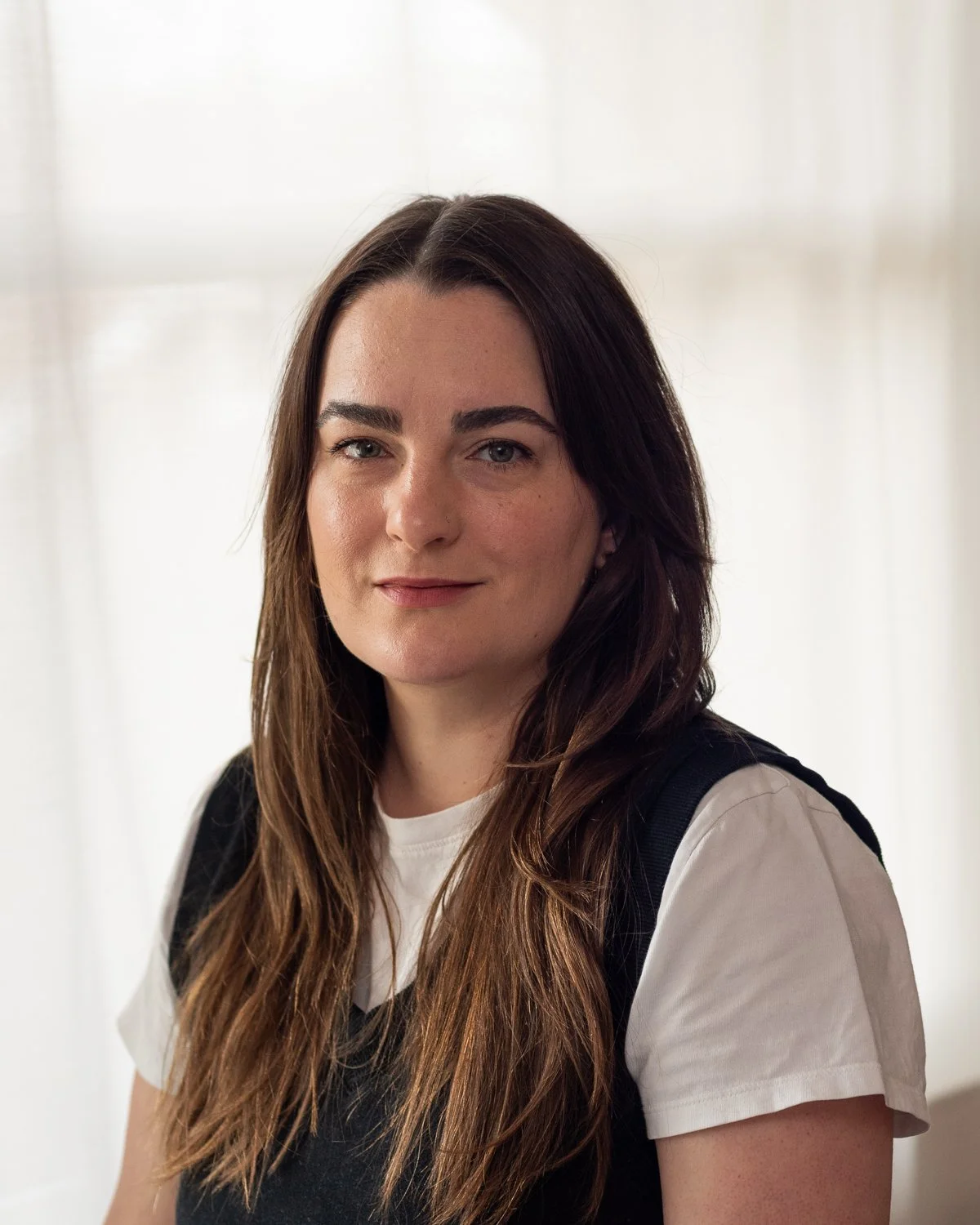 A young woman with long brown hair and fair skin, wearing a white t-shirt and a black backpack, standing in front of a light-colored background.