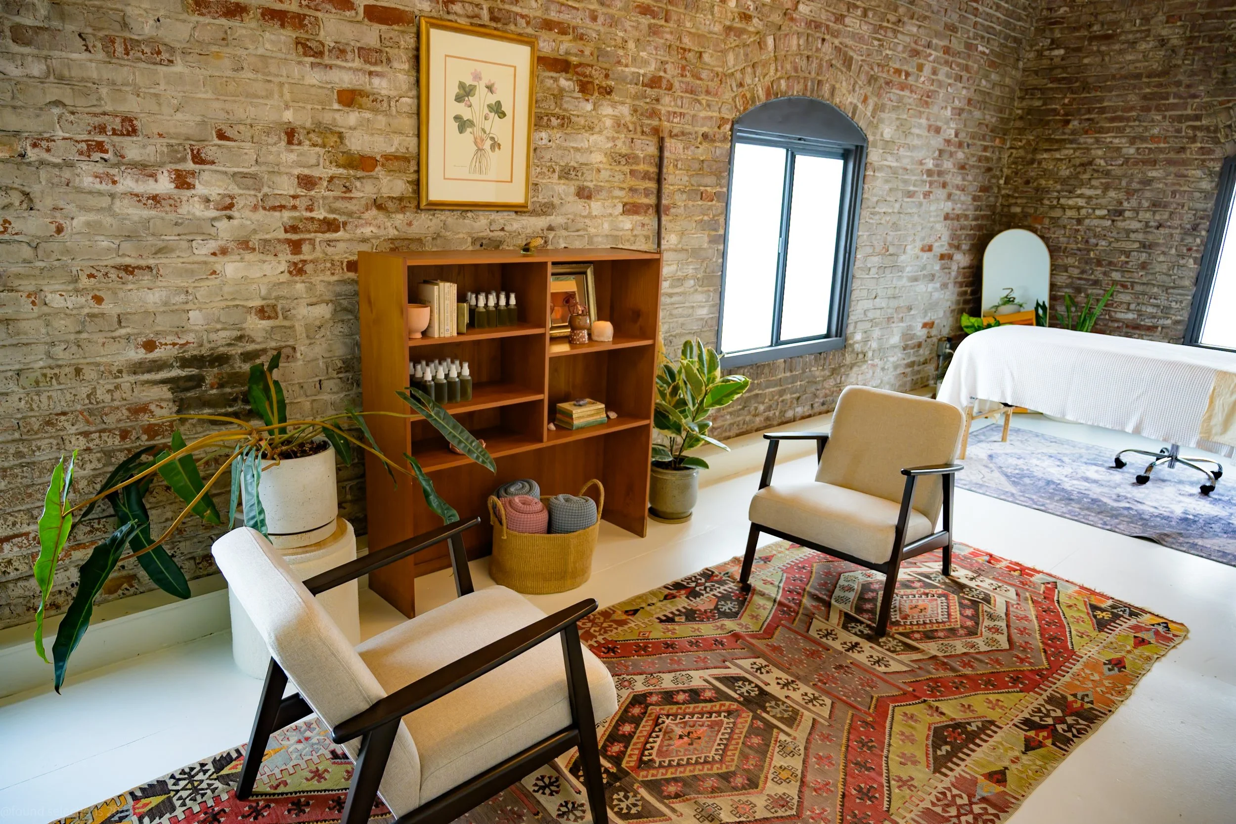 A cozy room with a rustic brick wall, wooden shelf with books and plants, two beige chairs, a red patterned rug, and a massage table in the background.
