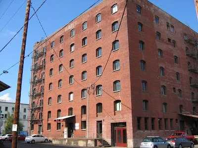 A multi-story red brick building with multiple arched and rectangular windows, located in an urban area with parked cars along the street.
