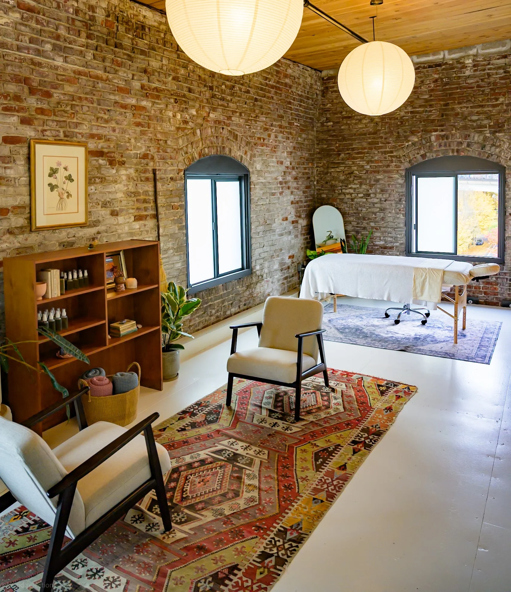 A light-filled massage therapy room with brick walls, a seating area, and healthy plants.
