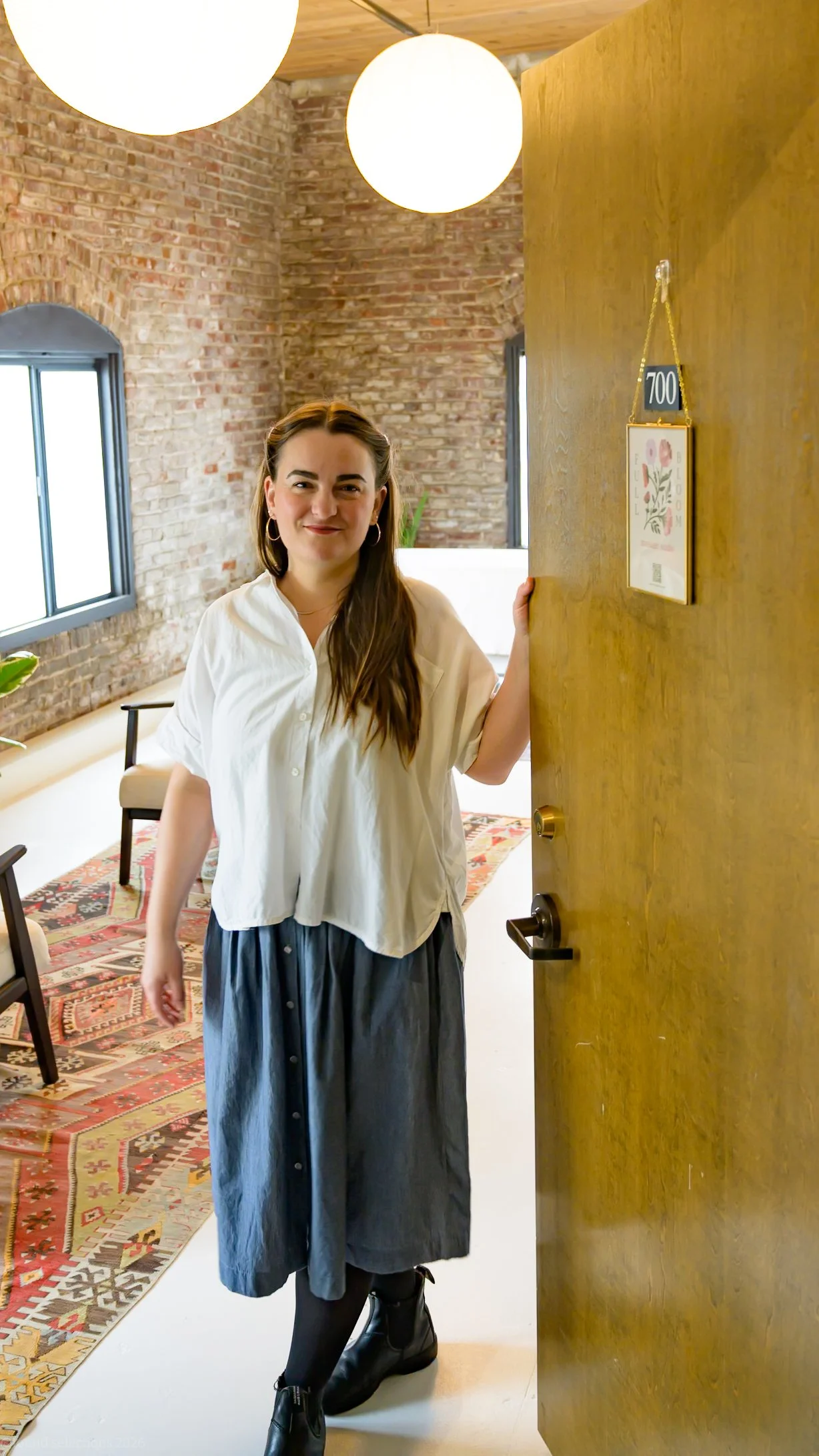 A woman with long brown hair, wearing a white blouse and a dark gray skirt, stands at the doorway of a cozy room with exposed brick walls and wooden ceiling beams, smiling at the camera.