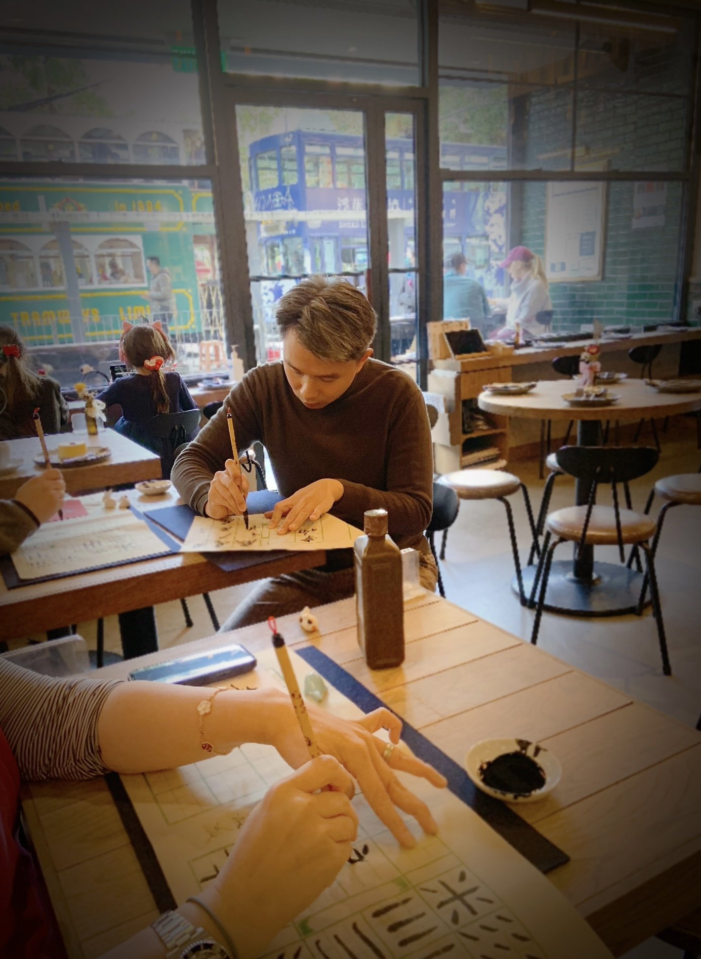 People practicing calligraphy at a cafe with a tram visible outside the window.