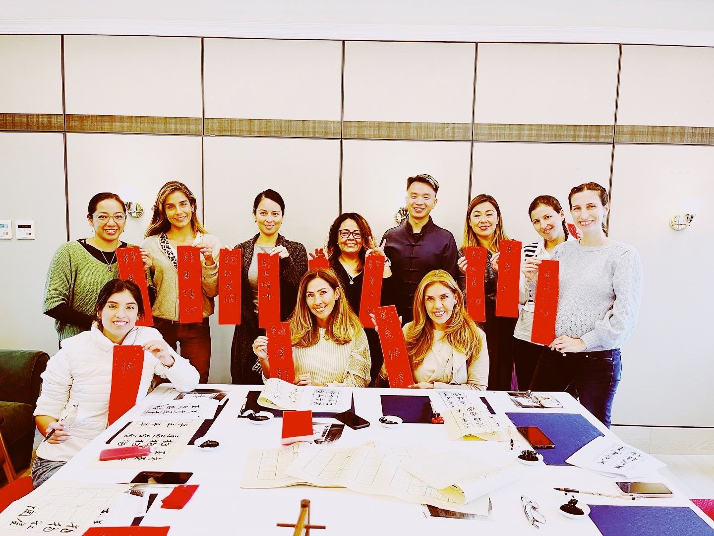 Group of people holding red papers with Chinese calligraphy, smiling and gathered around a table with writing materials.