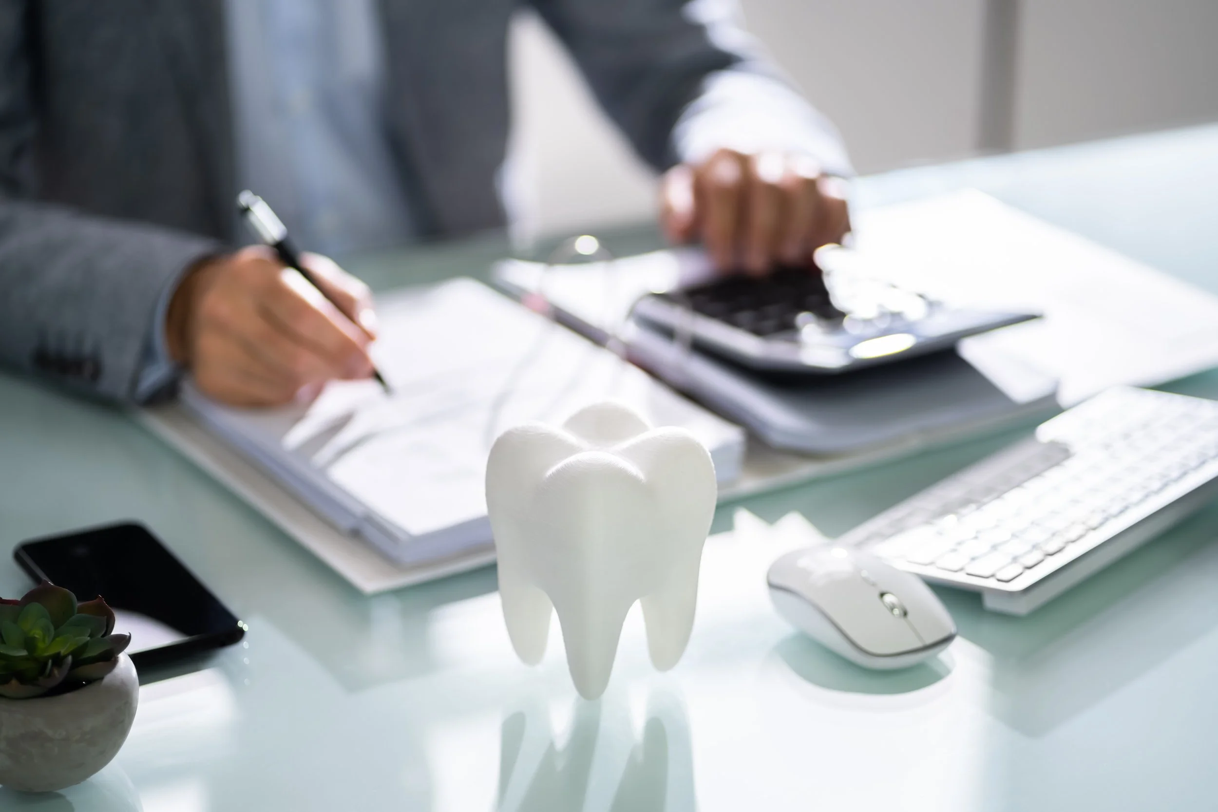 A model of a white tooth on a desk with a person writing and using a calculator in the background, along with a keyboard, mouse, smartphone, plant, and notepad.
