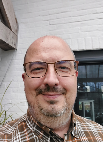 Close-up foto van een man met een glimlach, bril, met een baard en een kortgeknipt haar, gekleed in een geruit overhemd, voor een witte bakstenen muur en een raam.