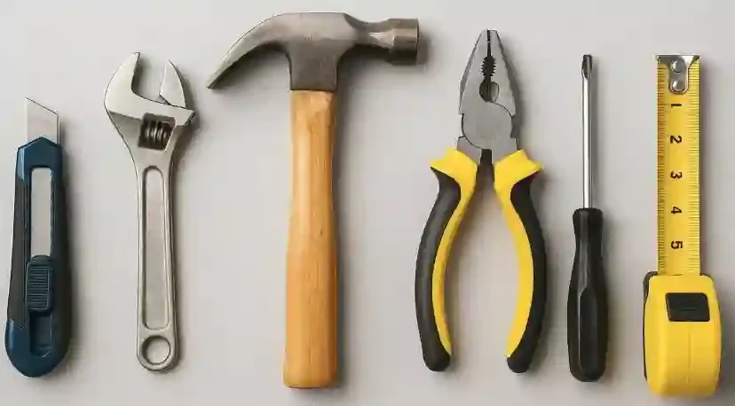 A row of six tools laid out on a white surface, including a utility knife, adjustable wrench, hammer, needle nose pliers, a small flathead screwdriver, and a tape measure.
