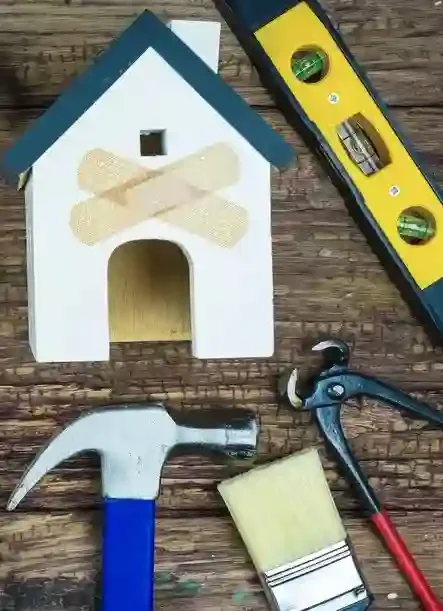 A small wooden doghouse with a blue roof, a level, a hammer, a paintbrush, and a pair of pliers on a wooden surface.