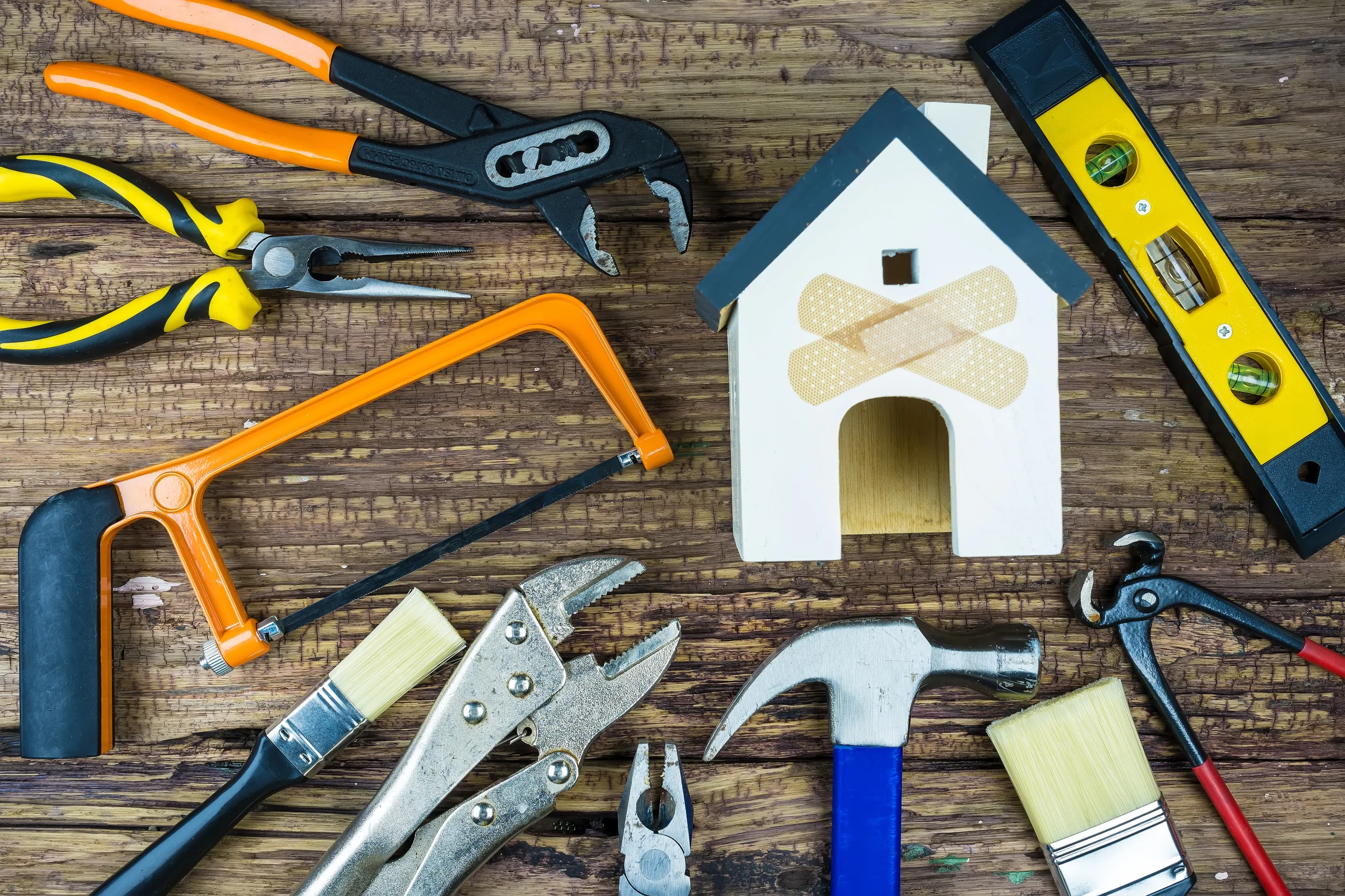 Tools and a small house model on a wooden surface, including pliers, a hacksaw, a hammer, a level, a handsaw, brushes, and a wrench.