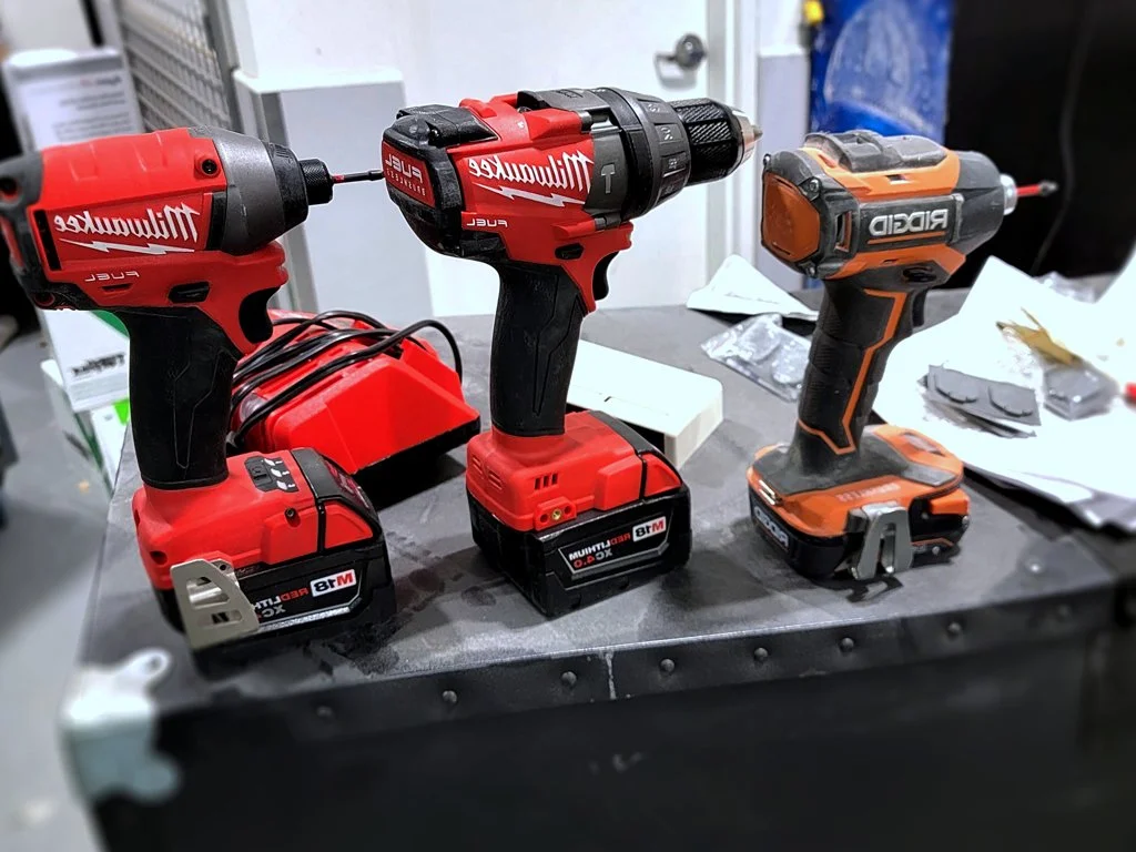 Two Milwaukee cordless power drills and one Ridgid cordless power drill on a workbench.