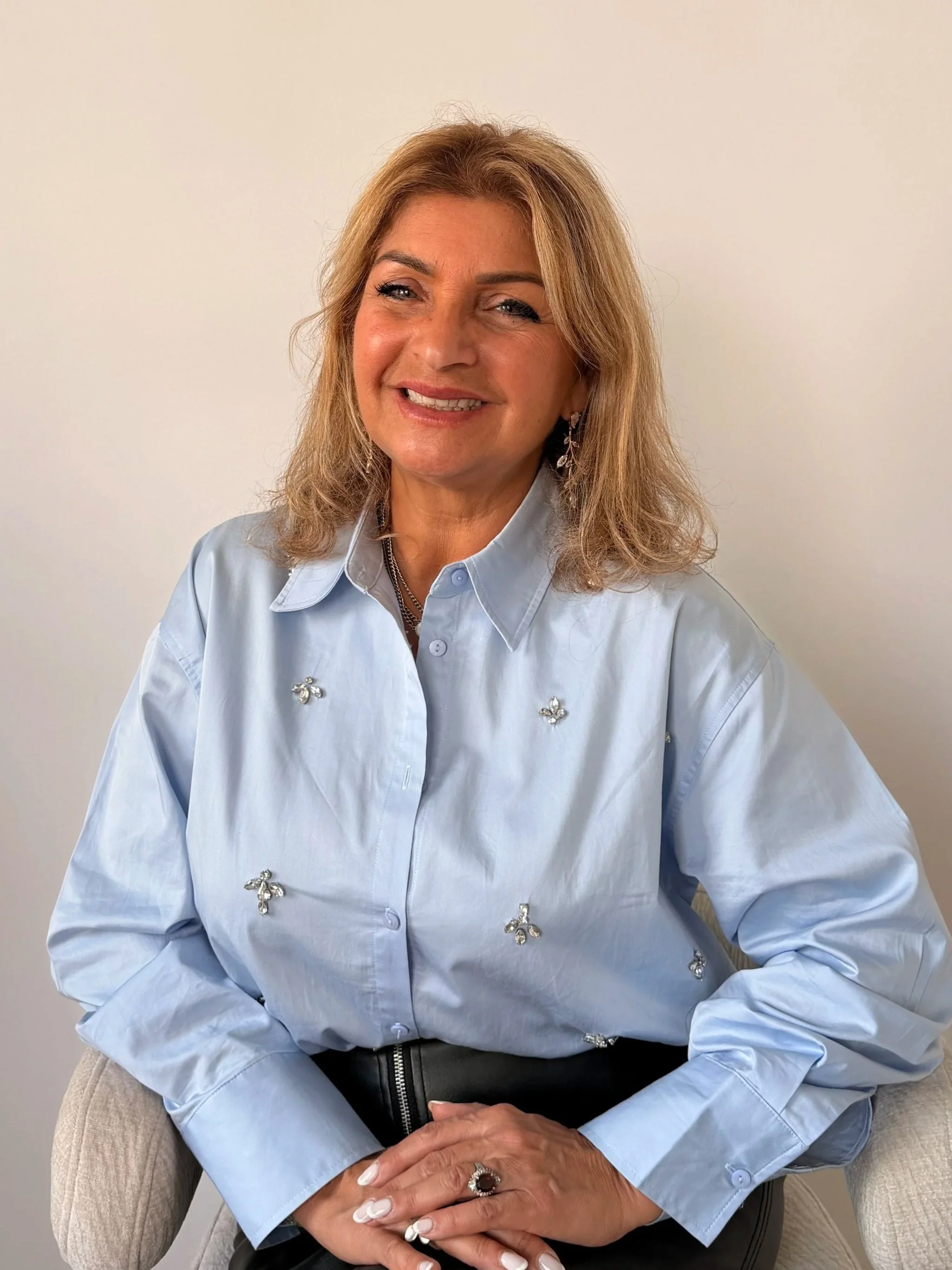 Une femme blonde souriante portant une chemise bleue ornée de broderies de petites abeilles, assise dans un fauteuil contre un mur blanc.