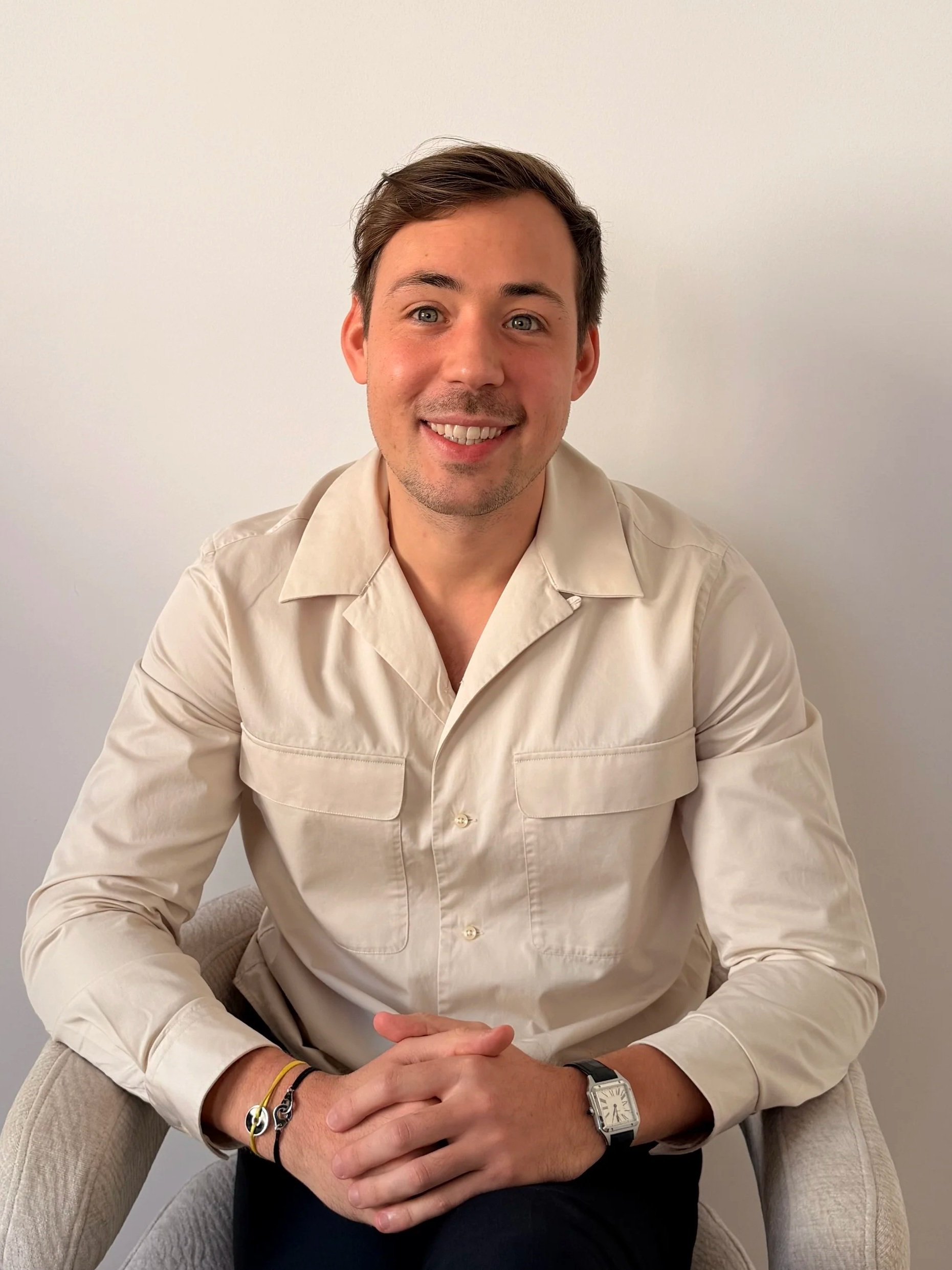 Un homme souriant, avec cheveux bruns, porte une chemise beige et une montre, assis sur un fauteuil contre un mur blanc.
