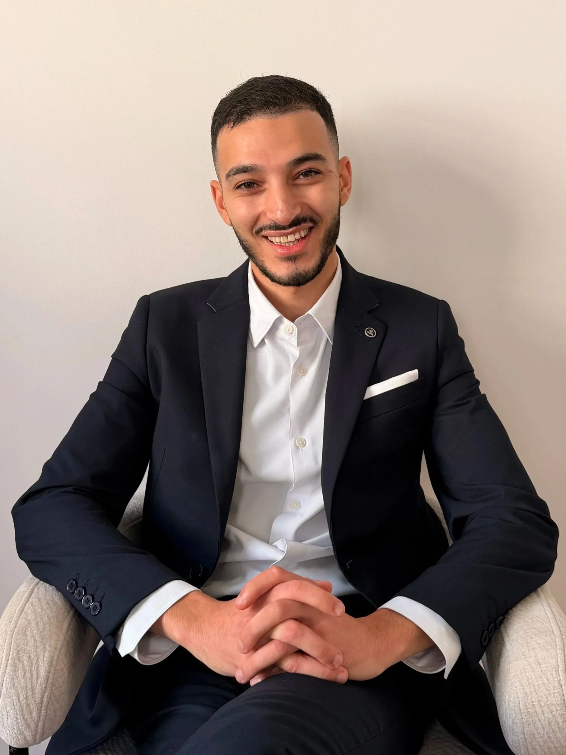 Jeune homme souriant, assis dans un fauteuil, portant un costume noir et une chemise blanche, fond blanc.