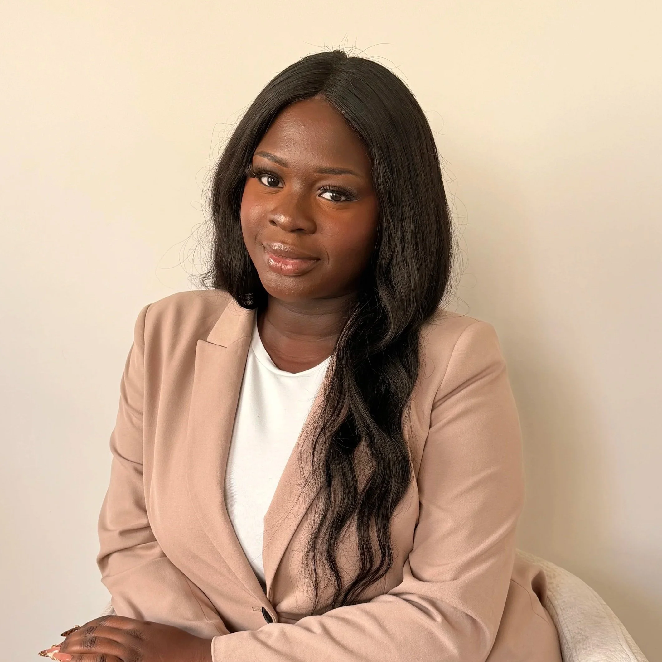 Une femme noire avec de longs cheveux noirs, portant une veste beige et un t-shirt blanc, assise contre un mur blanc.