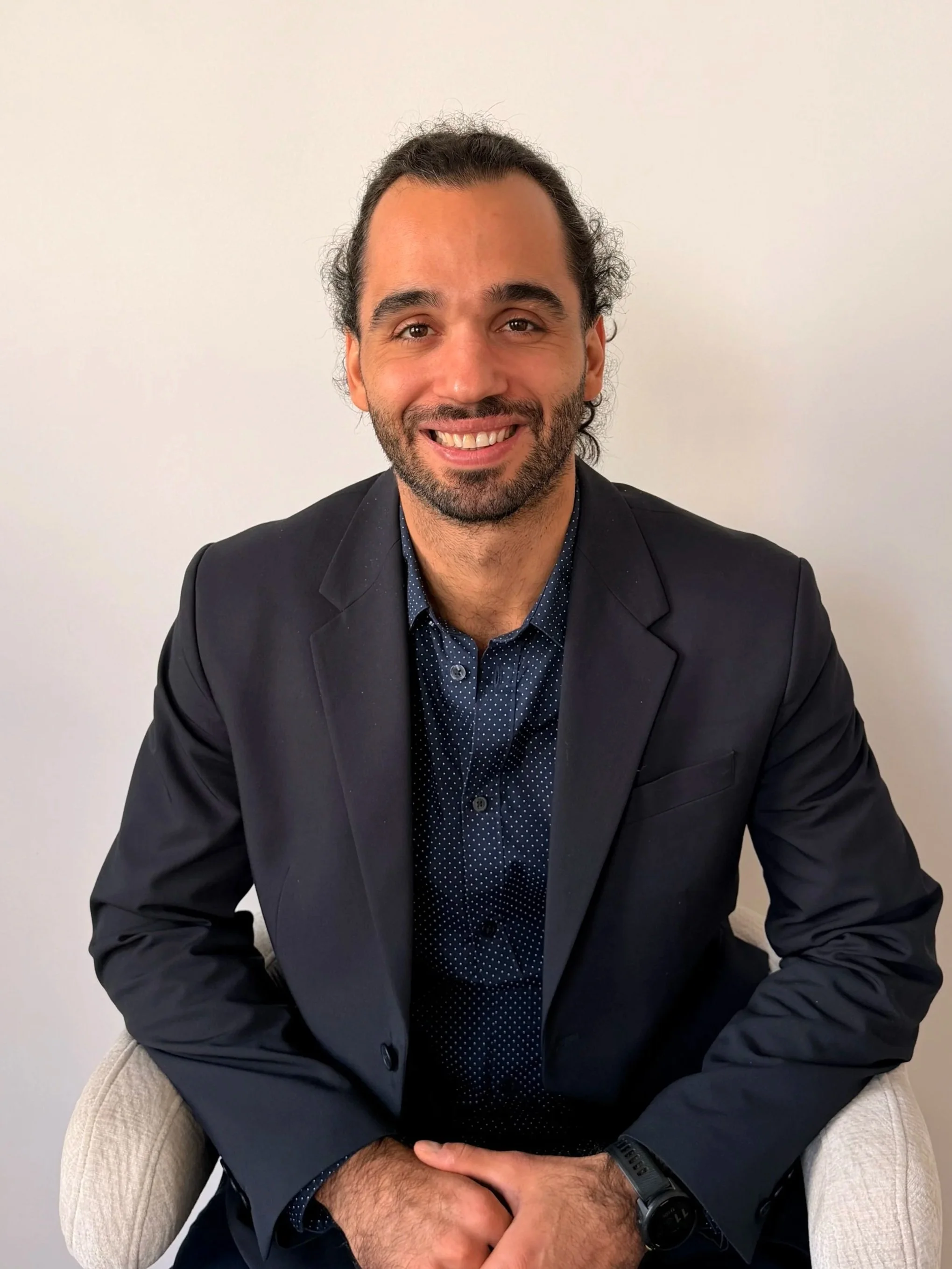 Un homme souriant habillé d'un costume noir, assis sur un fauteuil, contre un mur blanc.
