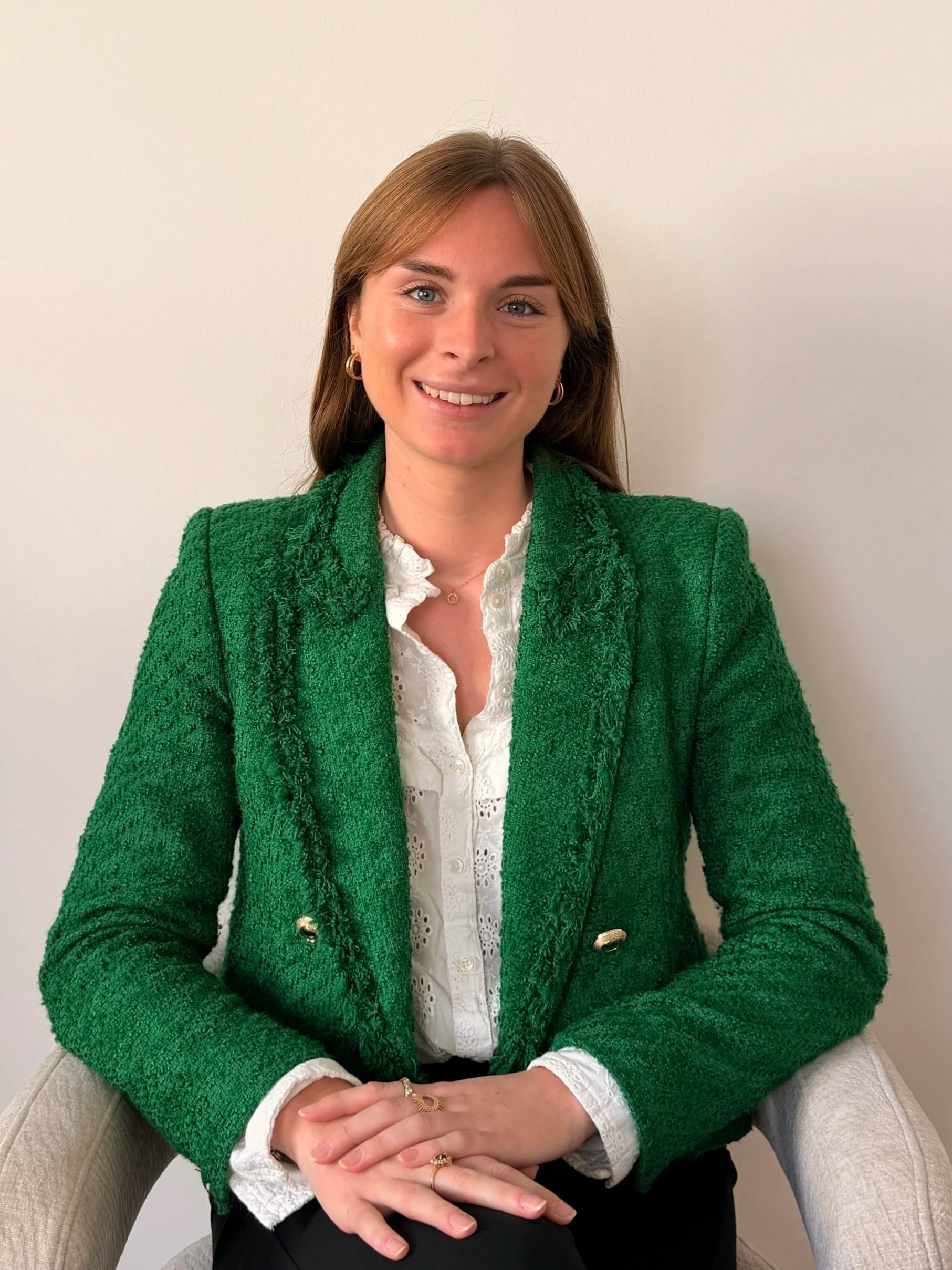 Femme souriante portant une veste verte assise sur un fauteuil beige devant un mur blanc