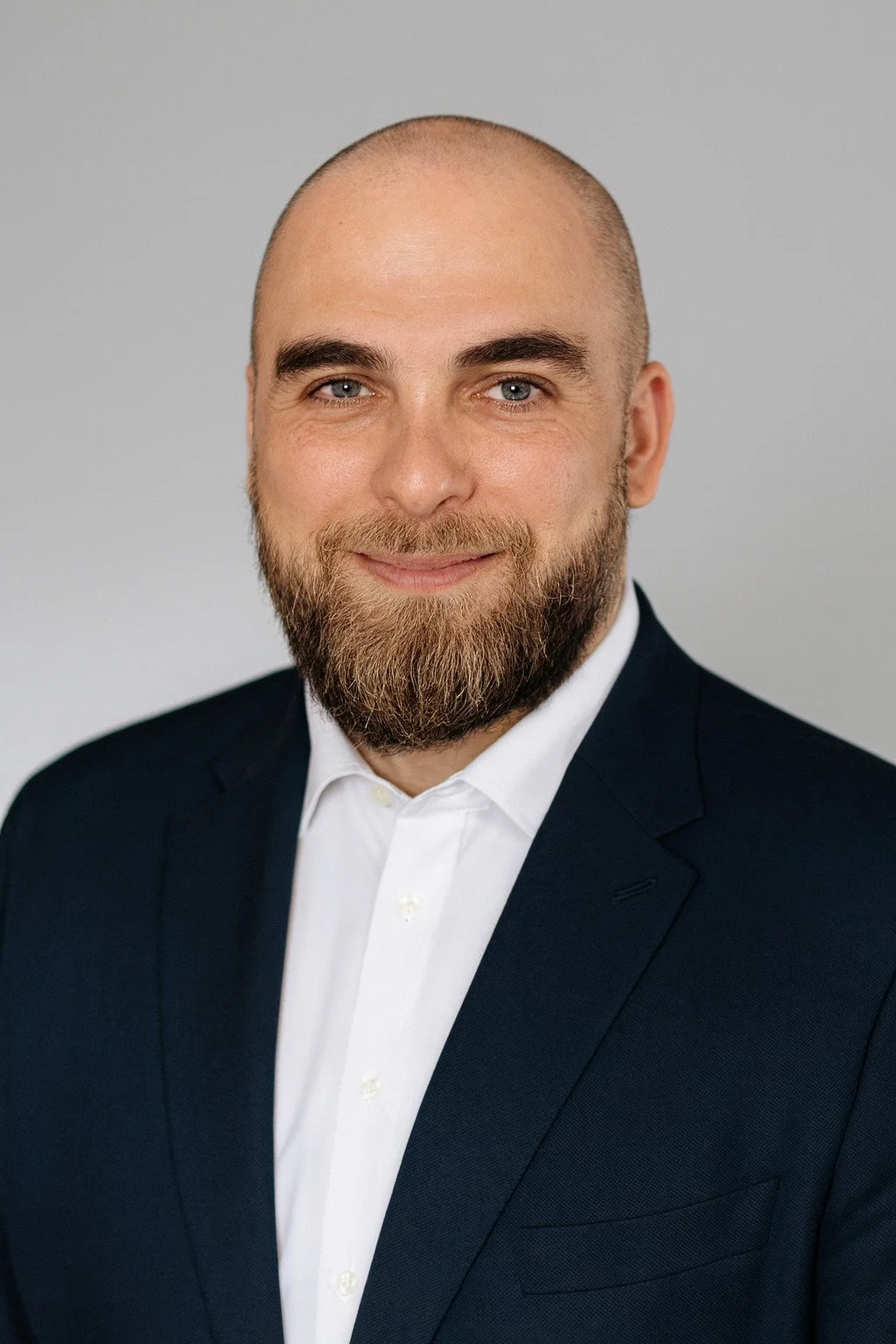 Un homme d'affaires souriant, portant un costume gris et une chemise blanche, assis devant un mur blanc.