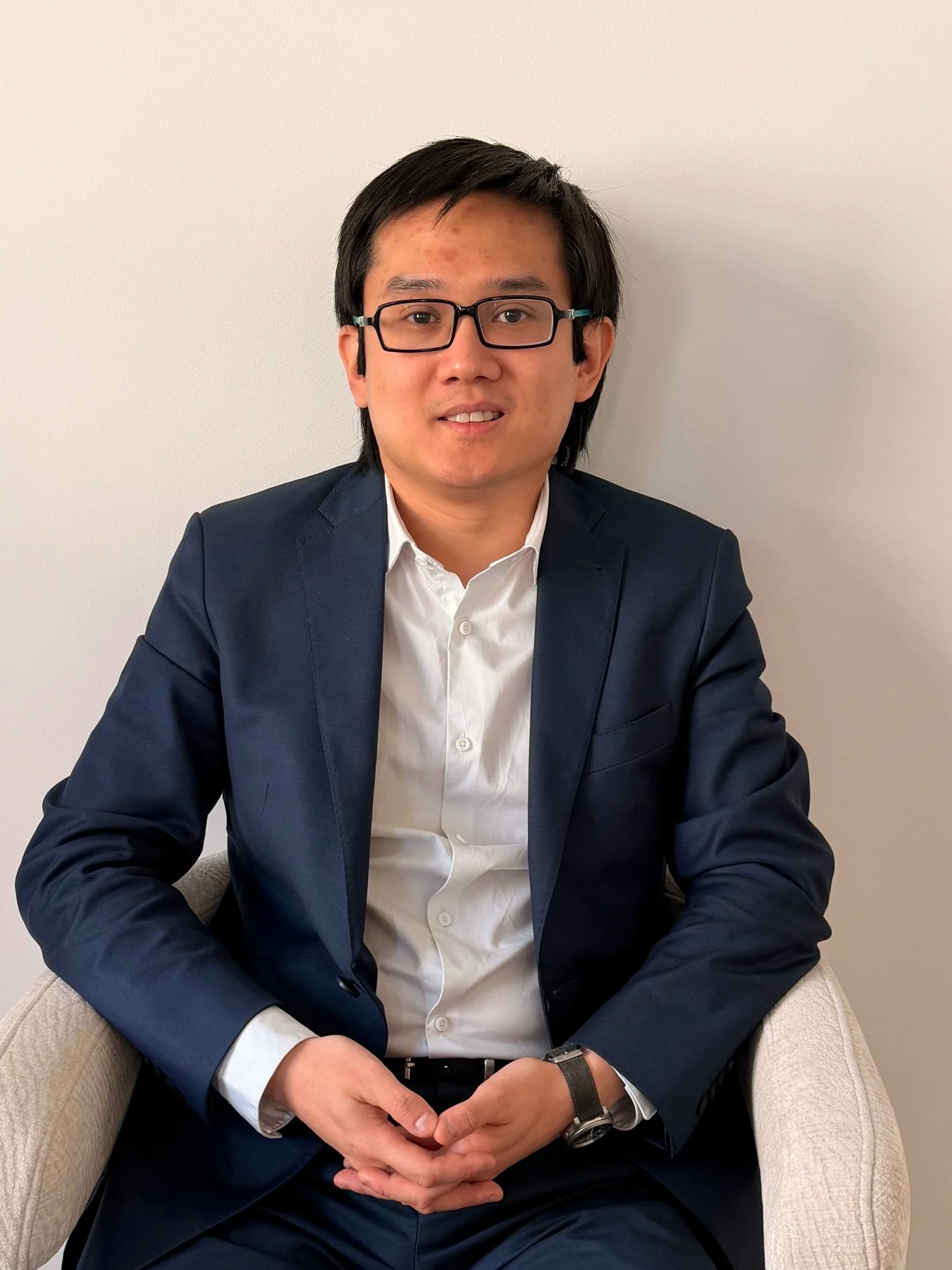 Un homme asiatique en costume noir et chemise blanche, portant des lunettes, assis sur un fauteuil beige contre un mur blanc.