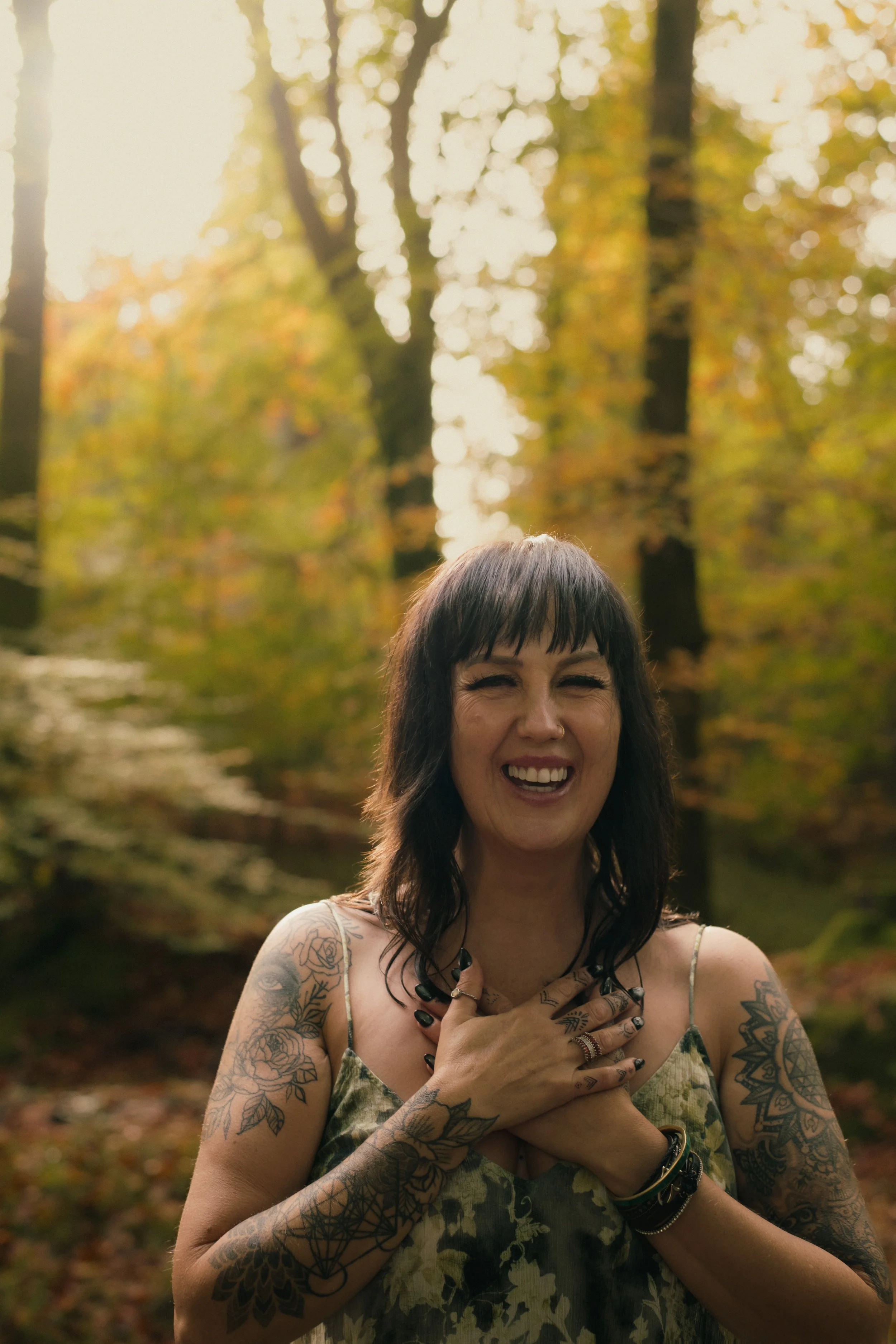 A woman with dark brown hair and tattoos on her arms, smiling and holding her hands over her chest, outdoors in a forest during autumn with trees and yellow leaves in the background.