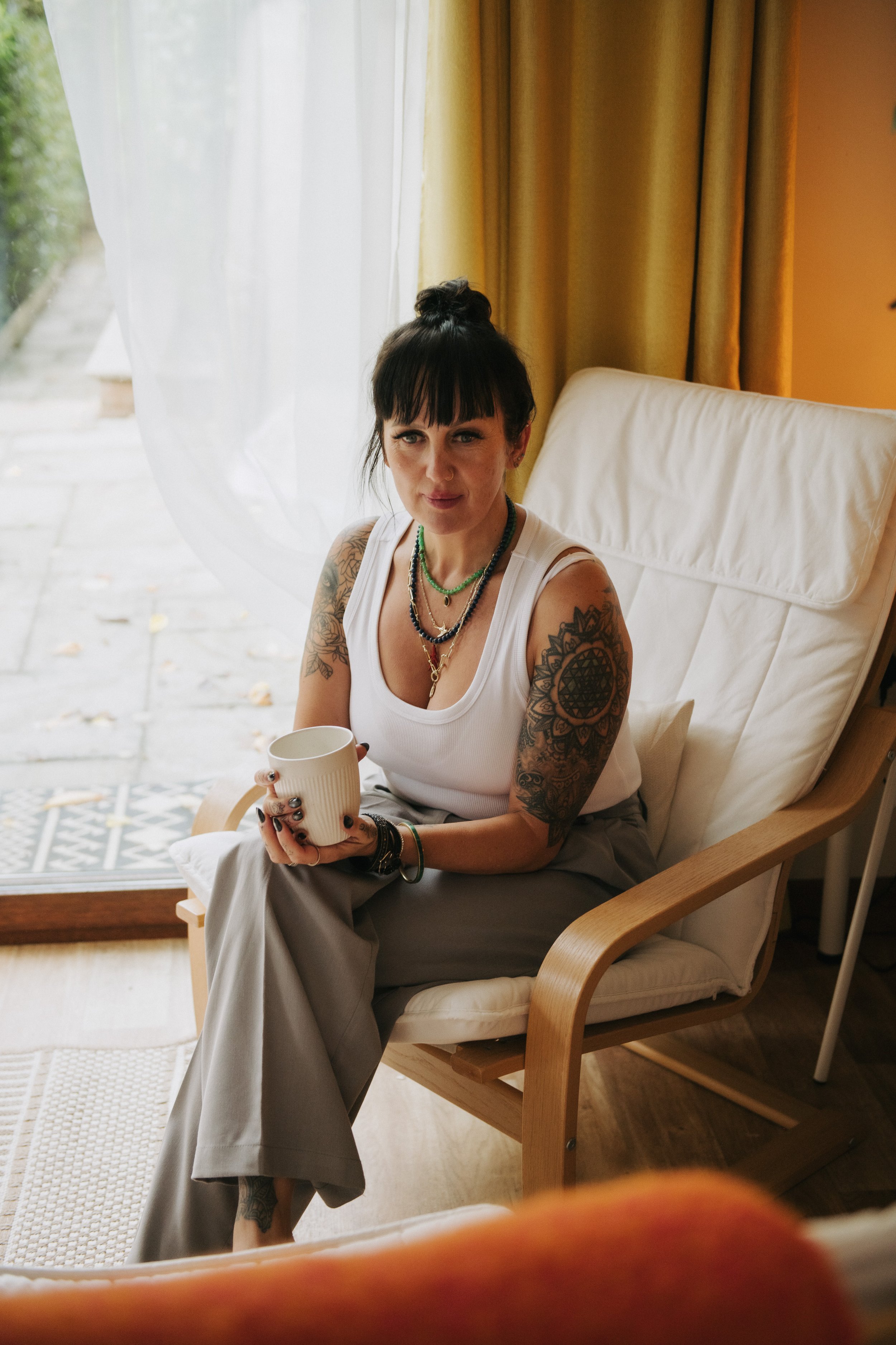 A woman with dark hair, tattoos on her arms, wearing a white tank top and light gray pants, is sitting in a wooden armchair holding a white mug. She is near a window with white curtains and yellow drapes, inside a cozy room with a wooden floor and warm lighting.