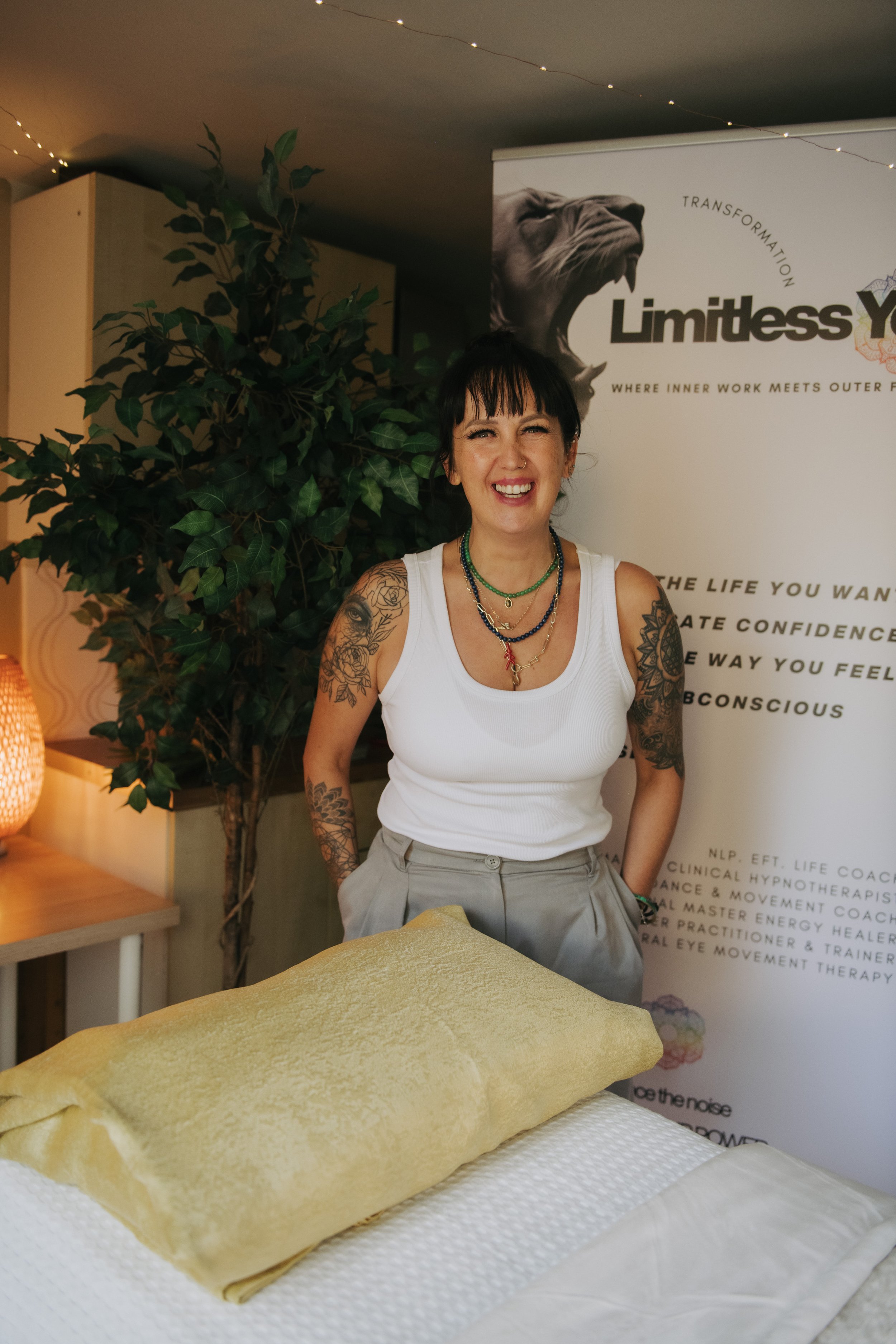 A woman with tattoos on her arms, wearing a white tank top and jewelry, smiling in front of a banner that reads 'Limitless You,' with a bed or massage table in the foreground.