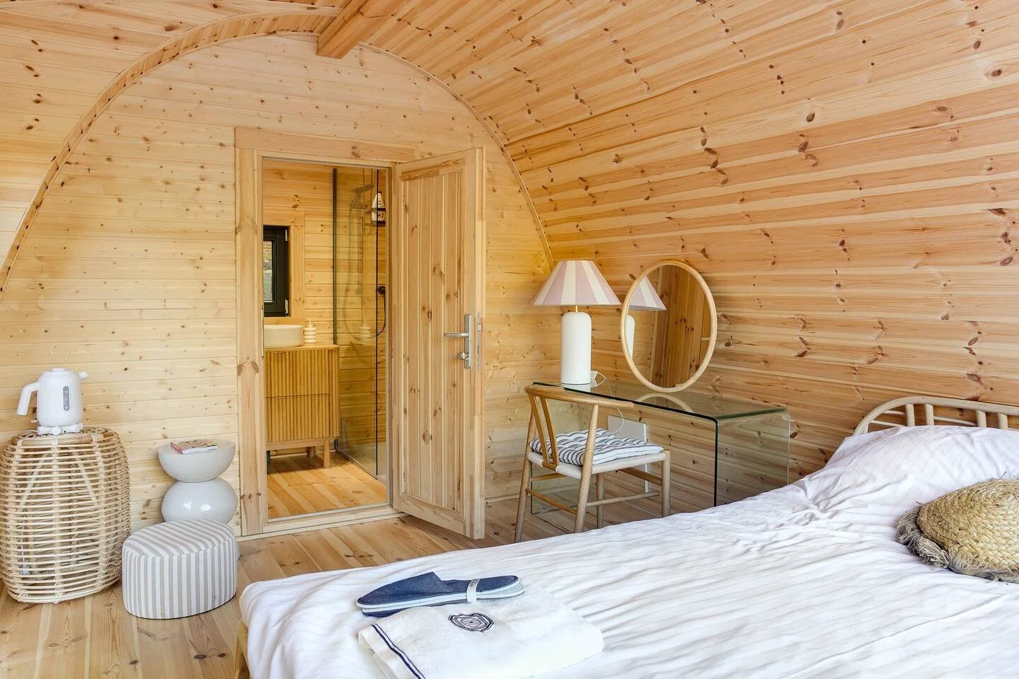 Houten slaapkamer met gewelfd plafond, een bed met wit beddengoed, glazen bureau met spiegel en lamp, en deur naar badkamer met douche.