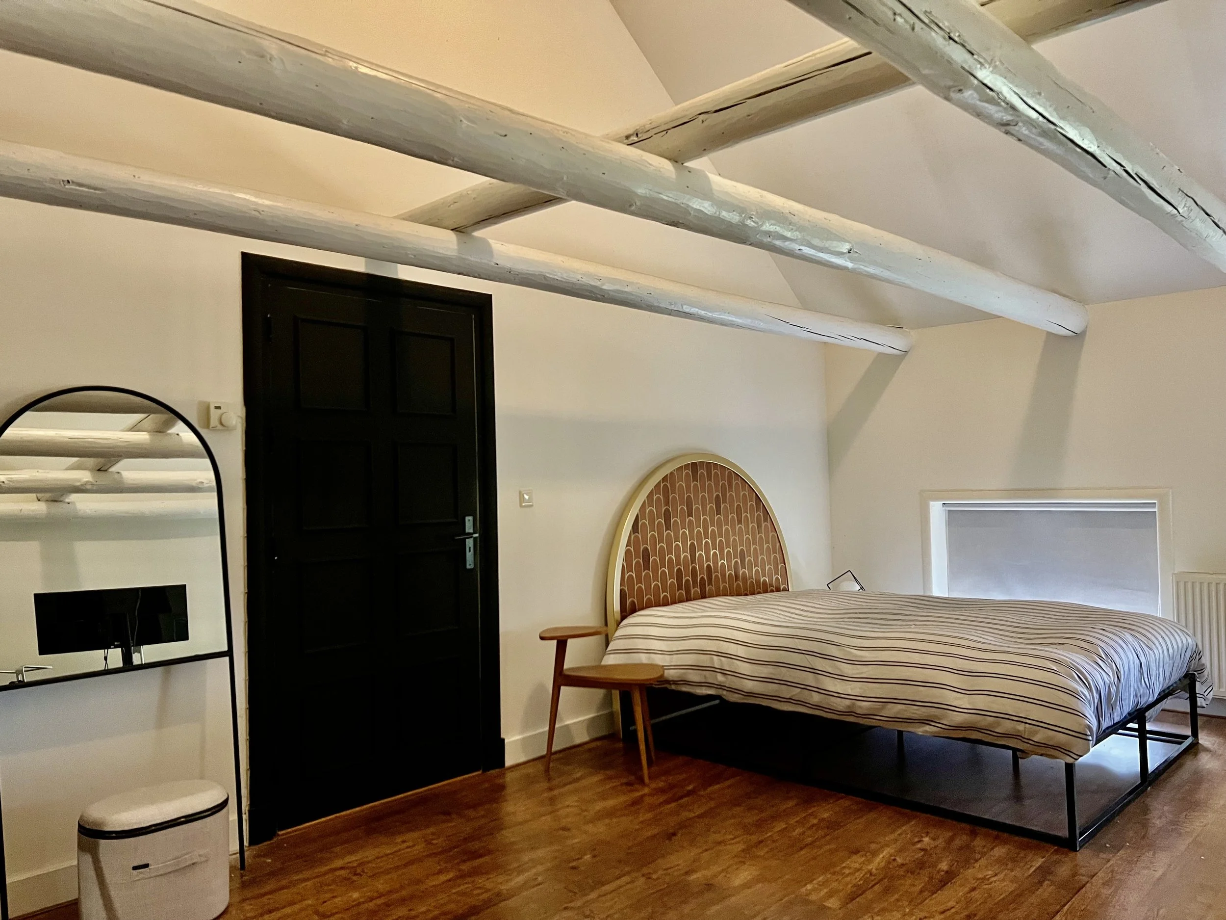 Slaapkamer met een tweepersoonsbed en gestreept dekbedovertrek, een kleine houten bijzettafel, houten vloer, gebogen spiegel naast een zwarte deur en zichtbare witte balken aan het plafond.