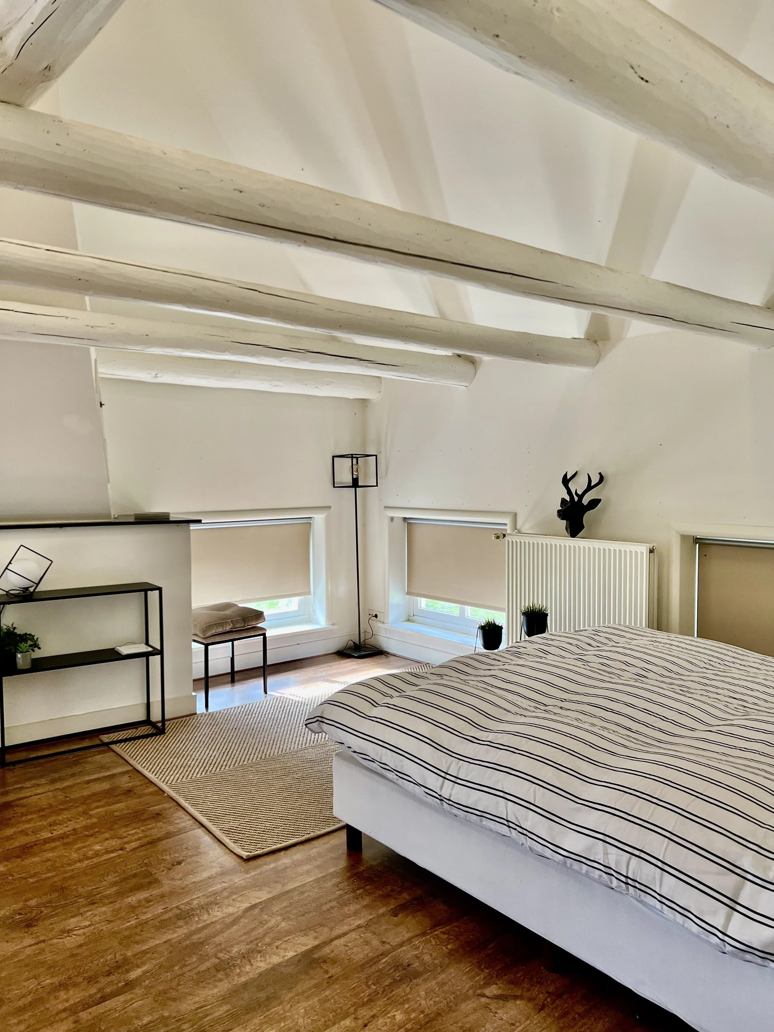 Slaapkamer met houten vloer, wit plafond met balken, bed met gestreept dekbedovertrek, raam met jaloezieën, planten, rek en decoratief hertengewei aan de muur.