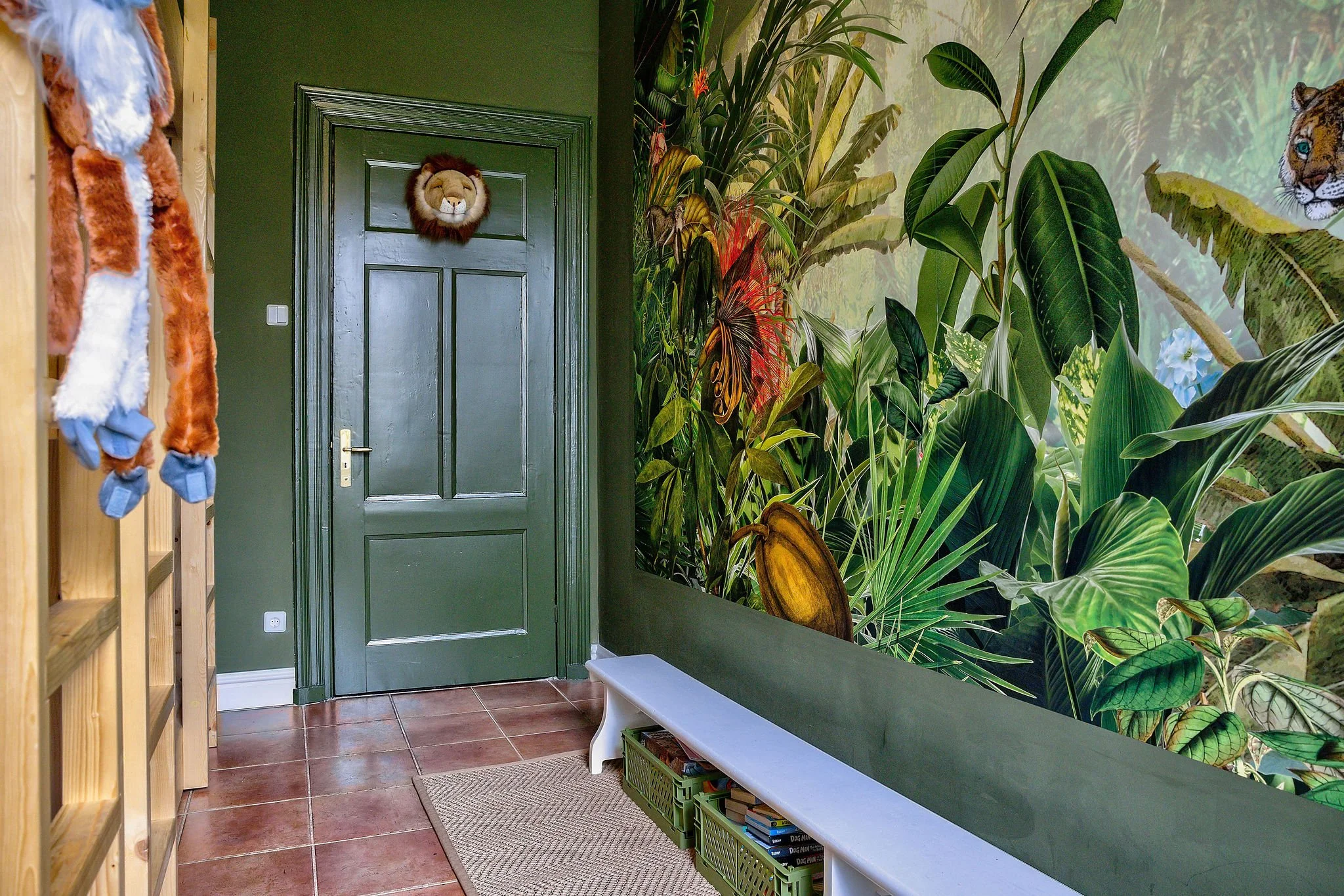 Groene kamer met deur versierd met leeuwenhoofd, jungle-muurschildering, stoffen aap aan houten meubel, bankje met manden en boeken.