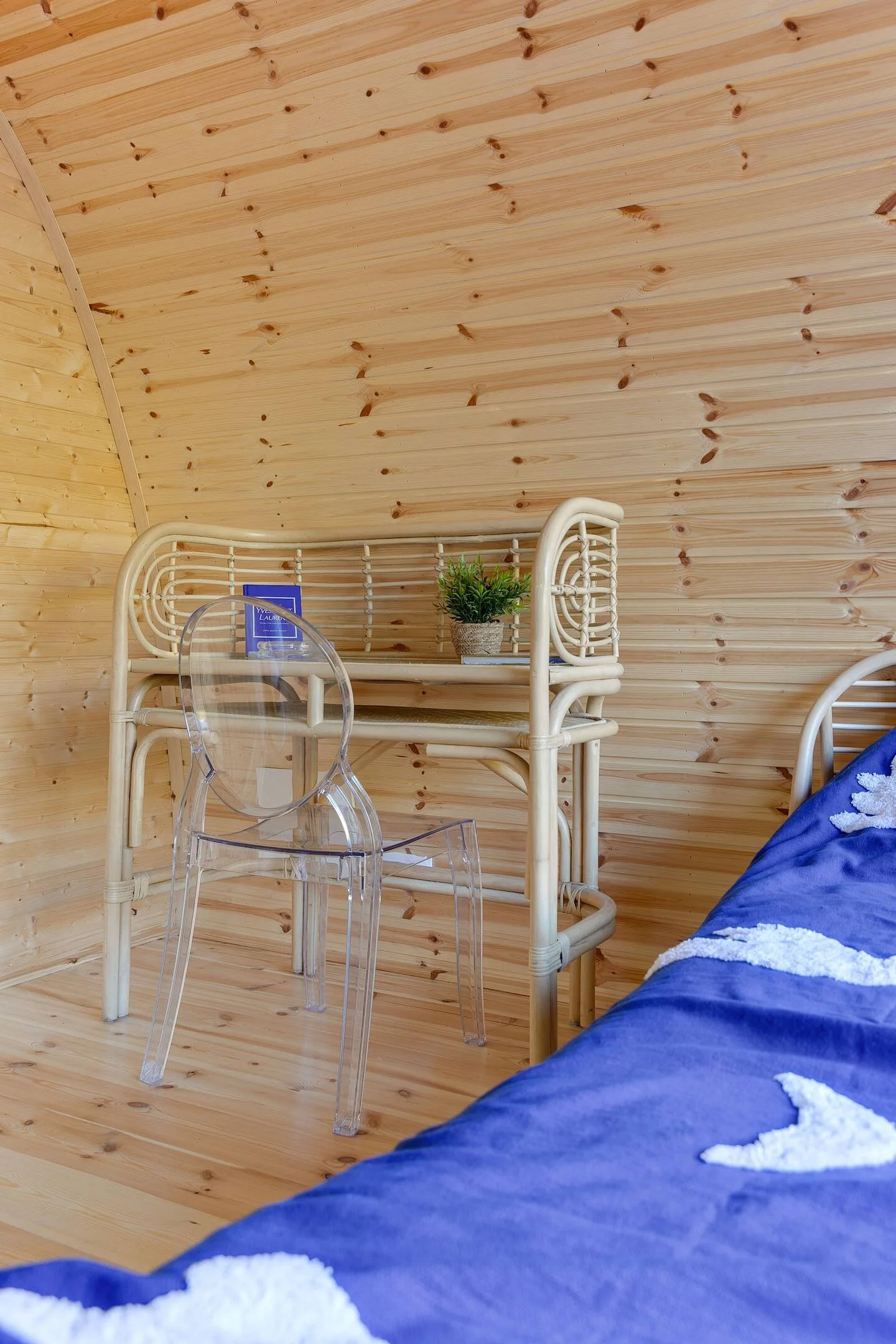 Interieur van een houten kamer met een rotan bureau, een doorzichtige stoel en een klein plantje op het bureau. Er is een blauw beddegoed zichtbaar.