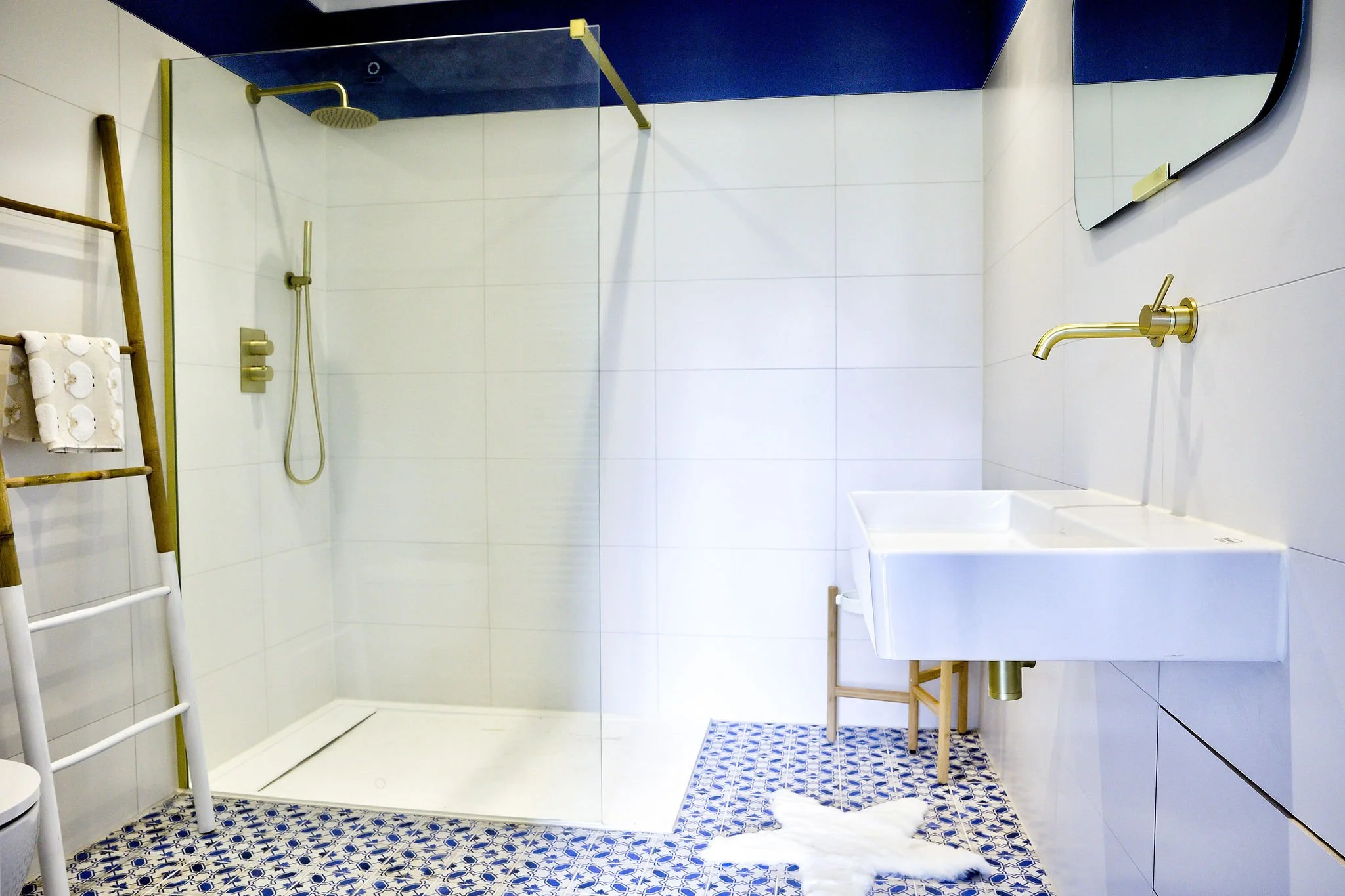 Moderne badkamer met glazen douchewand, gouden douchekop en kraan, witte tegels en blauw-witte patroontegels op de vloer.