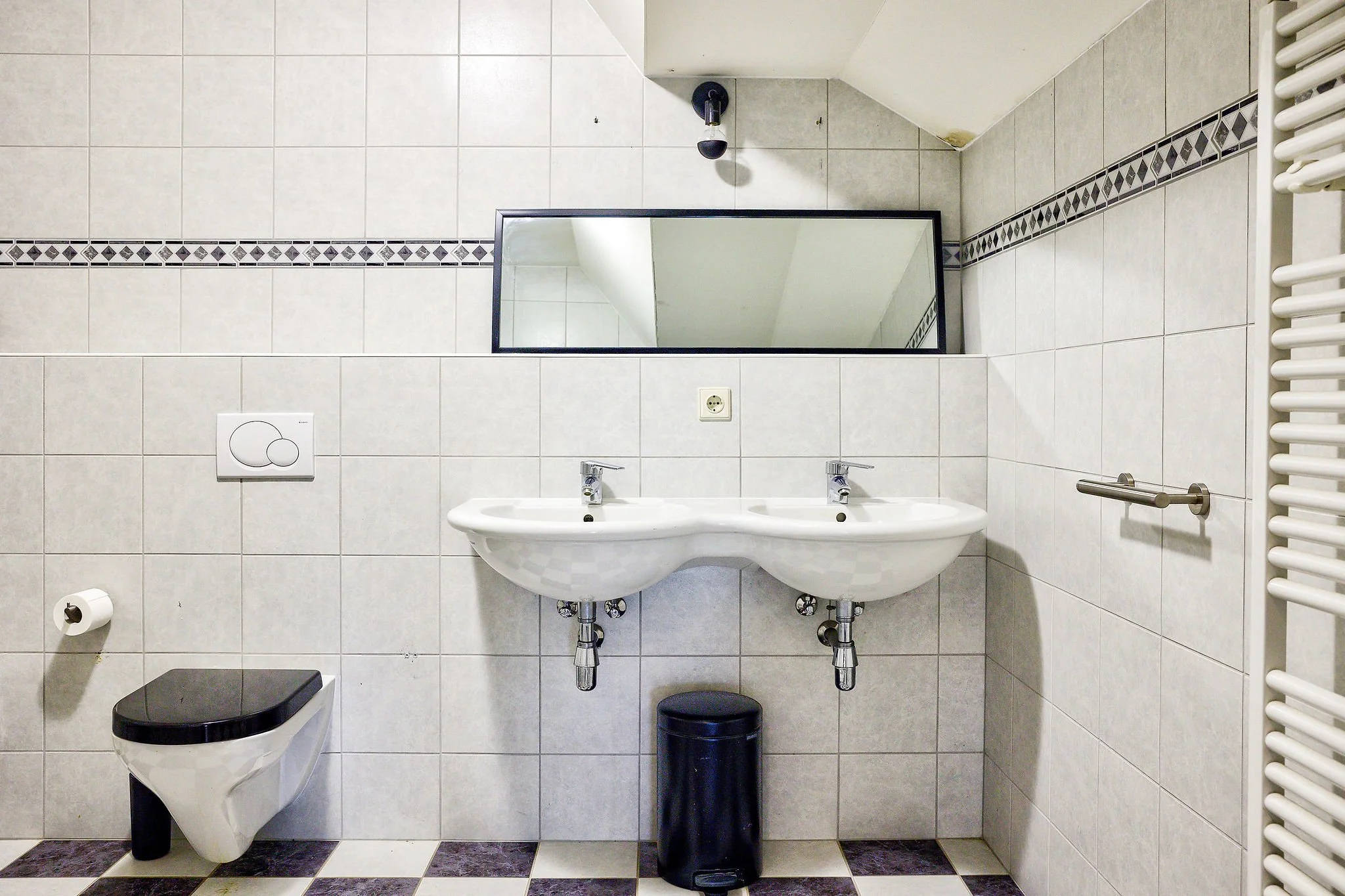 Badkamer met toilet, dubbele wastafel, spiegel, toiletpapierhouder, radiator, en prullenbak.