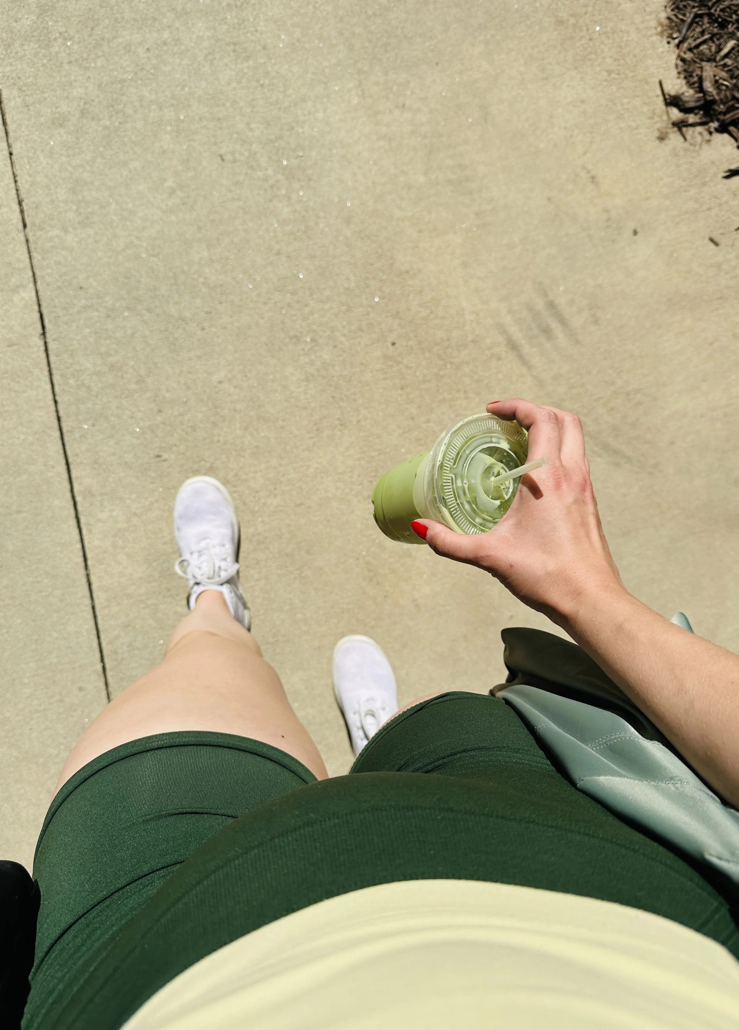 Photo prise du point de vue d'une personne marchant sur un trottoir, portant un short vert, des chaussures de sport blanches, tenant une boisson vert clair à la main.