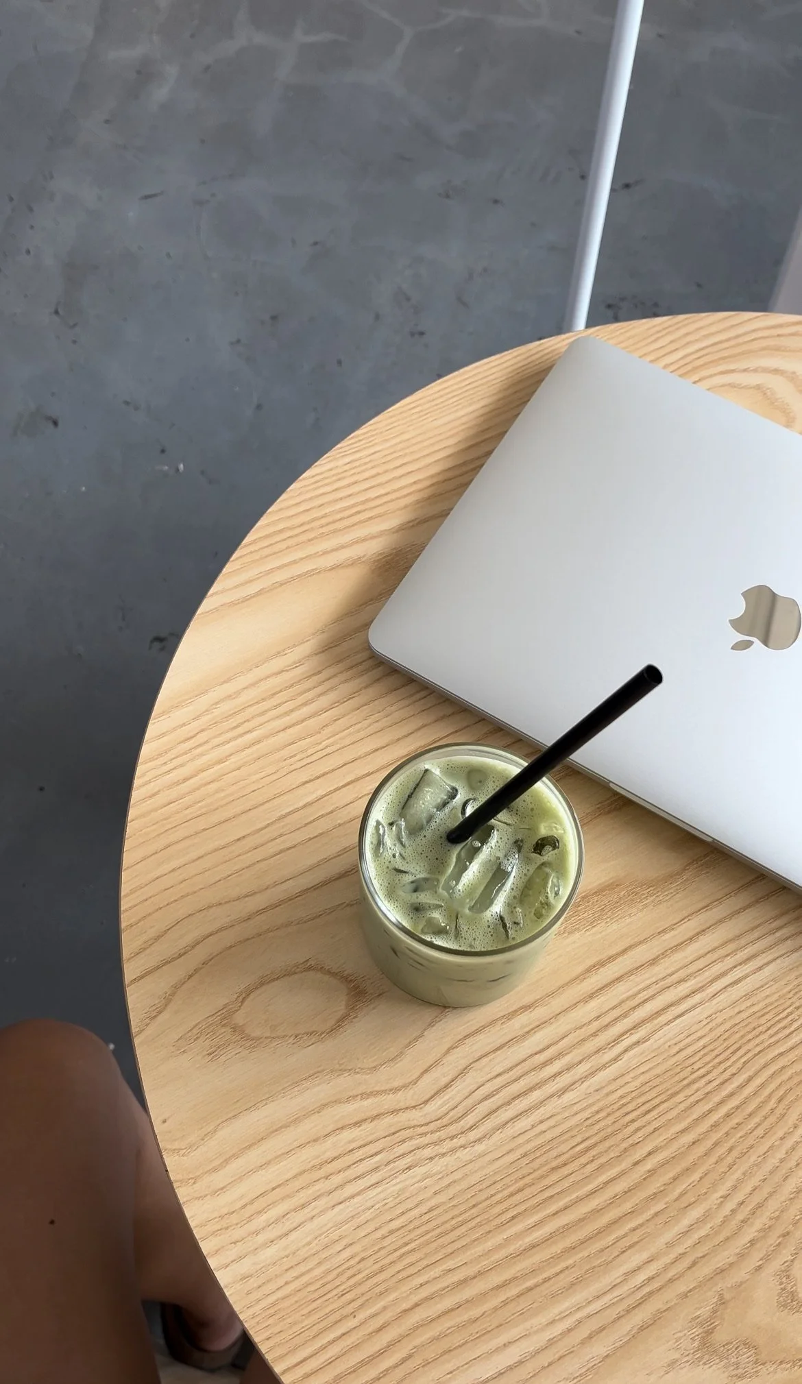 Un verre de matcha avec glaçons et un paille noir, une ordinateur portable blanc Apple, posé sur une table en bois clair.