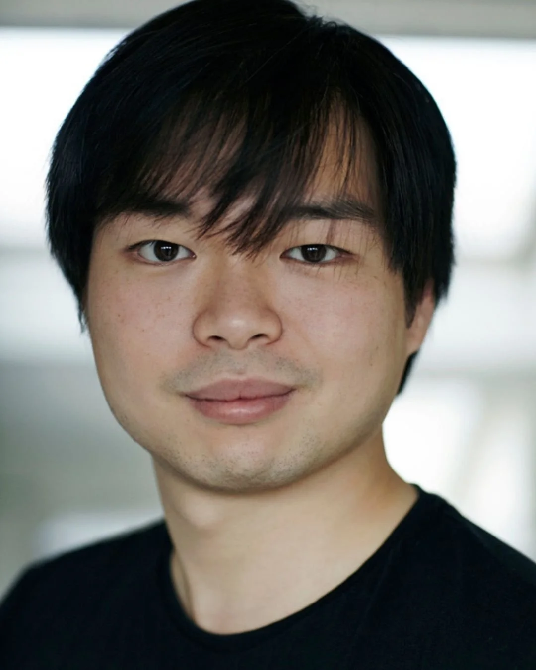 Close-up portrait of a young Asian man with dark hair and brown eyes, smiling slightly, wearing a black shirt.