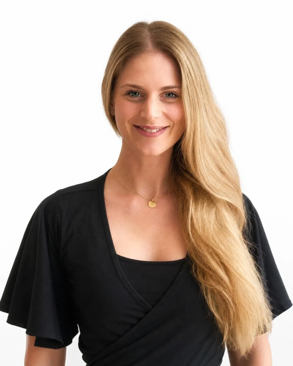 A woman with long blonde hair smiling, wearing a black top and a gold necklace with a small pendant, standing against a plain white wall.